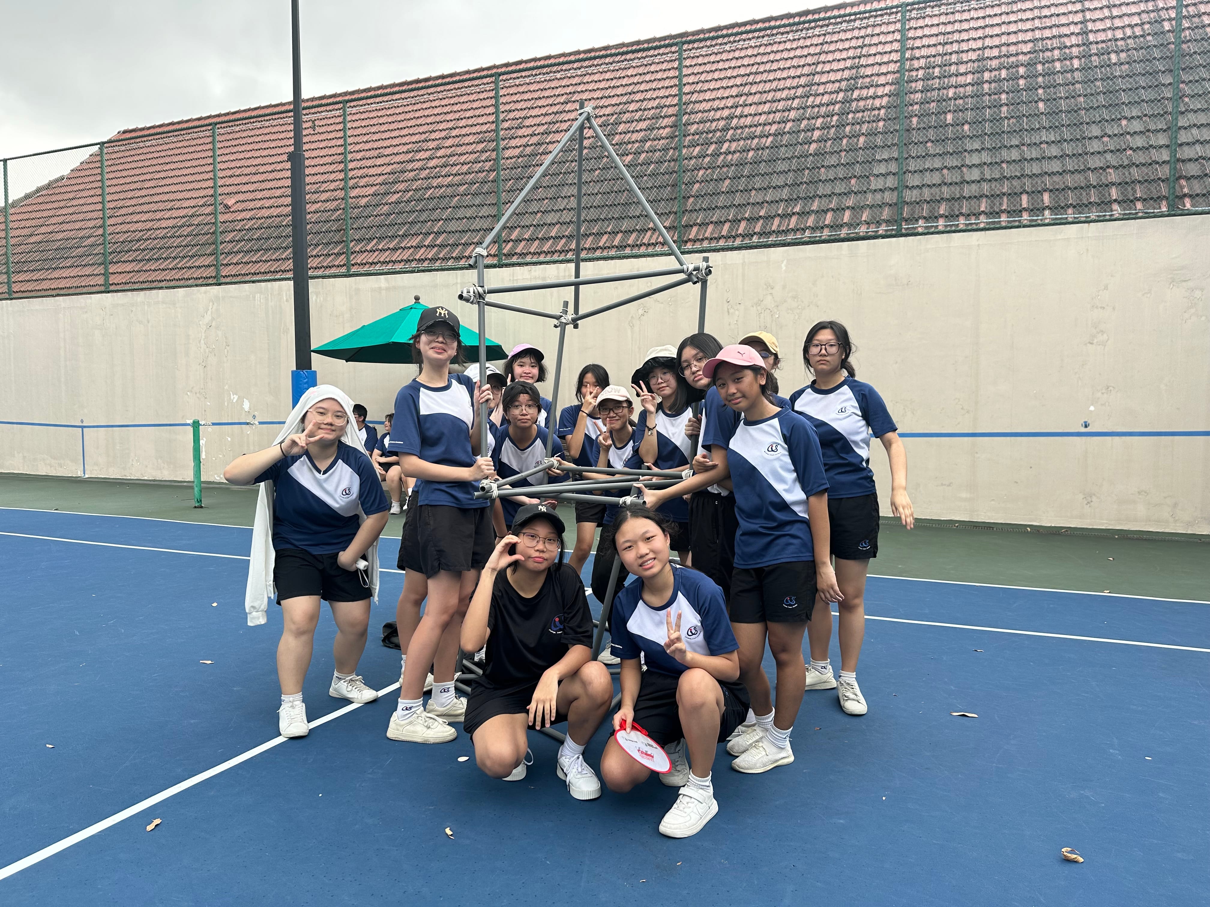 A group of young students in navy blue uniforms posing together on a blue tennis court, holding a metal frame structure.