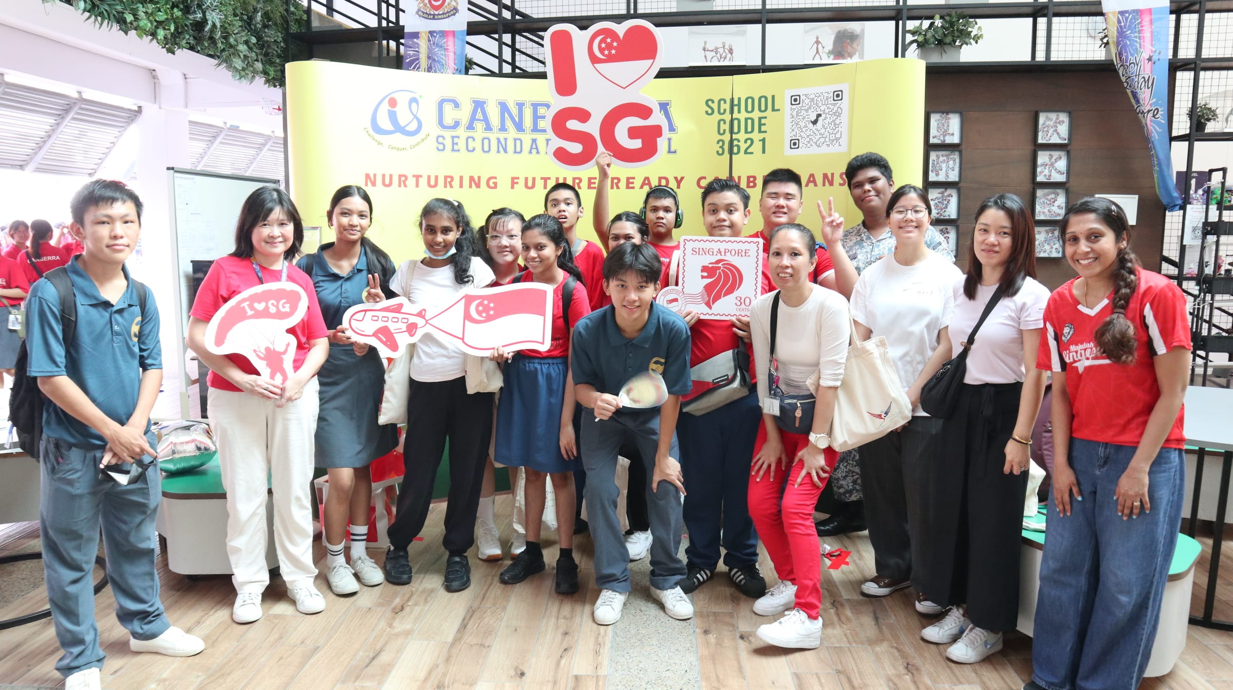 Group of students and adults posing in front of a Canesia Secondary School banner with I Love Singapore props.
