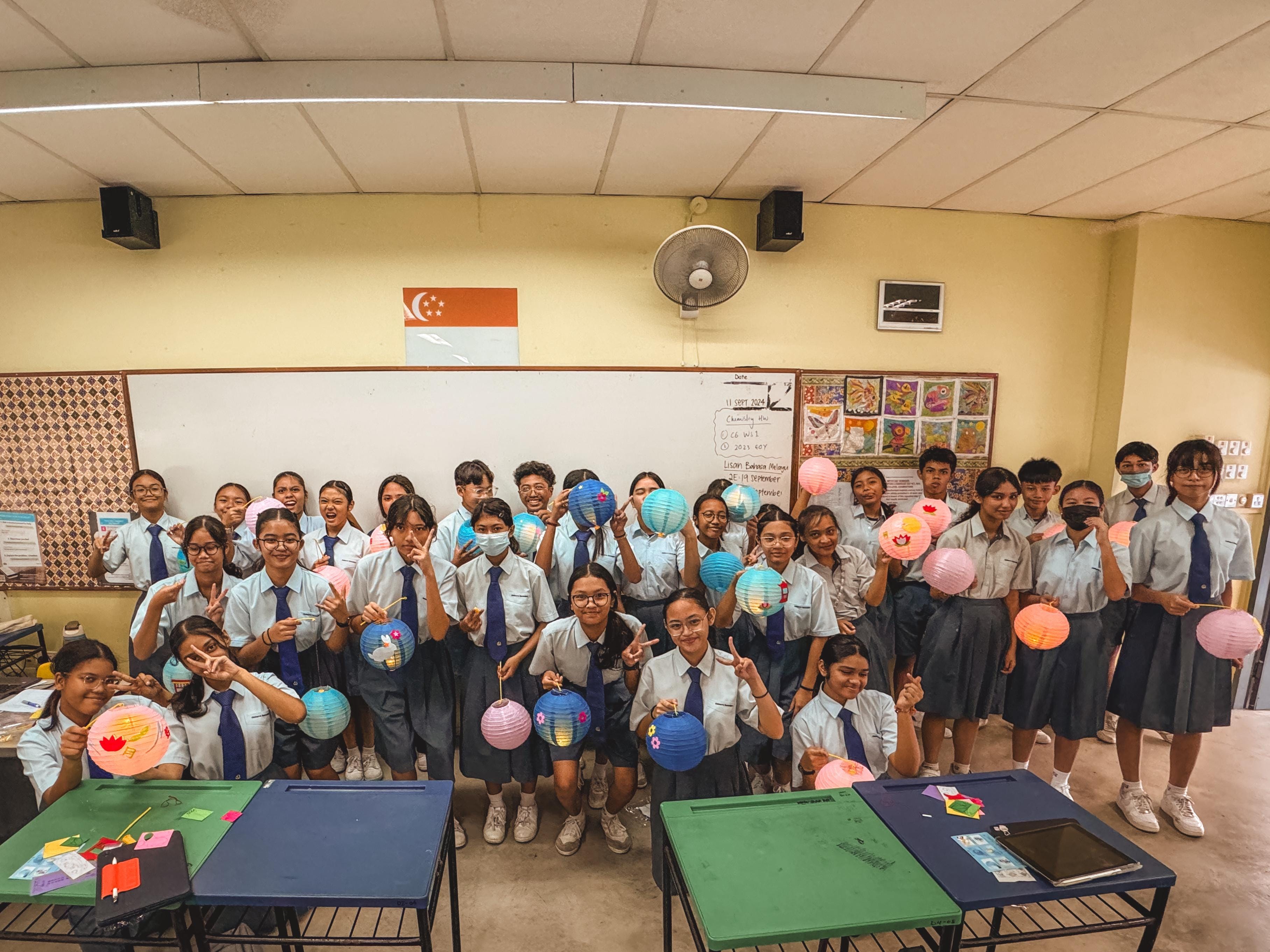 A large group of school students in uniforms holding colorful paper lanterns in a classroom with a Singapore flag.