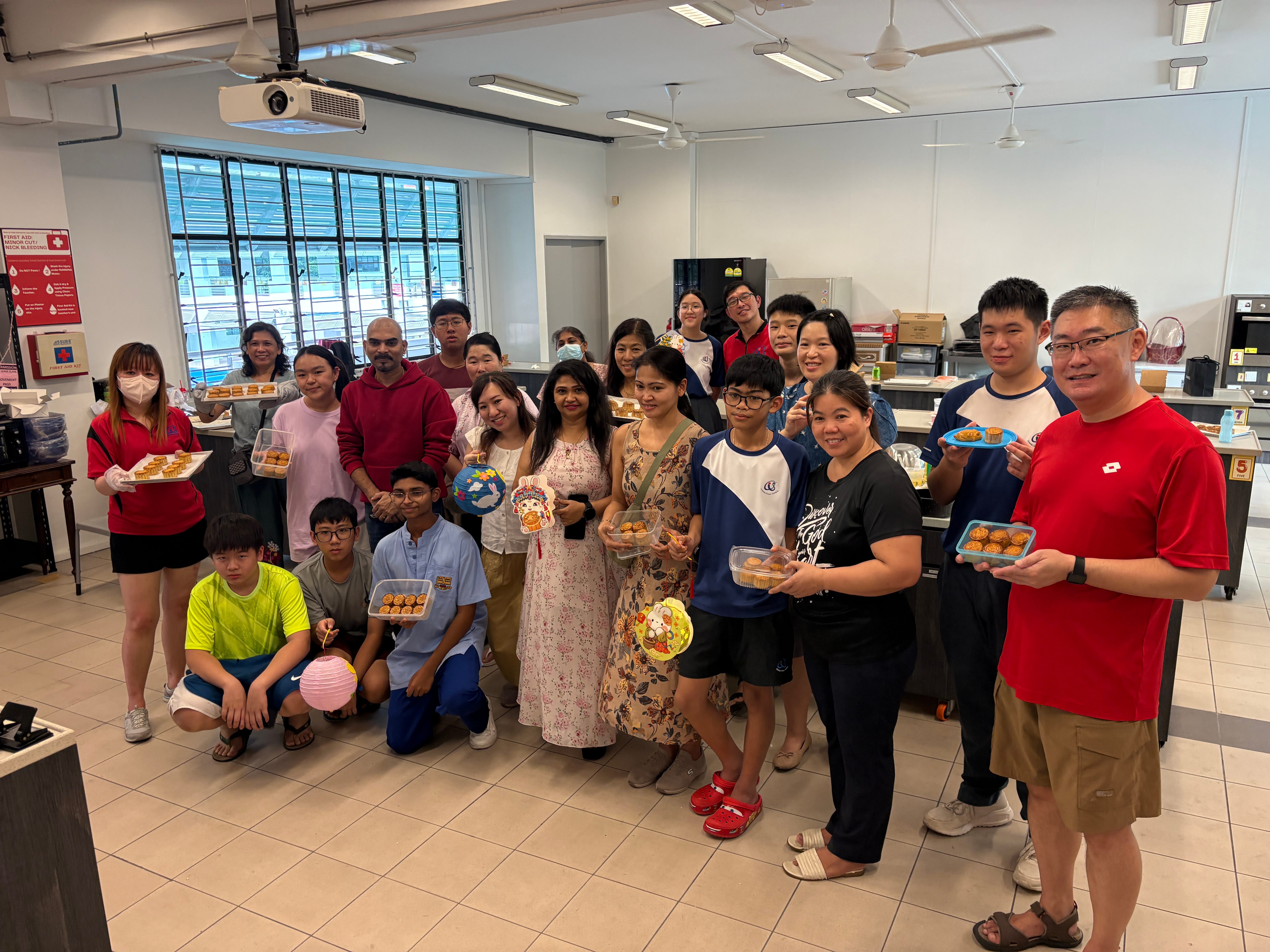 Group of people in a classroom kitchen posing with trays of pastries and colorful lanterns.