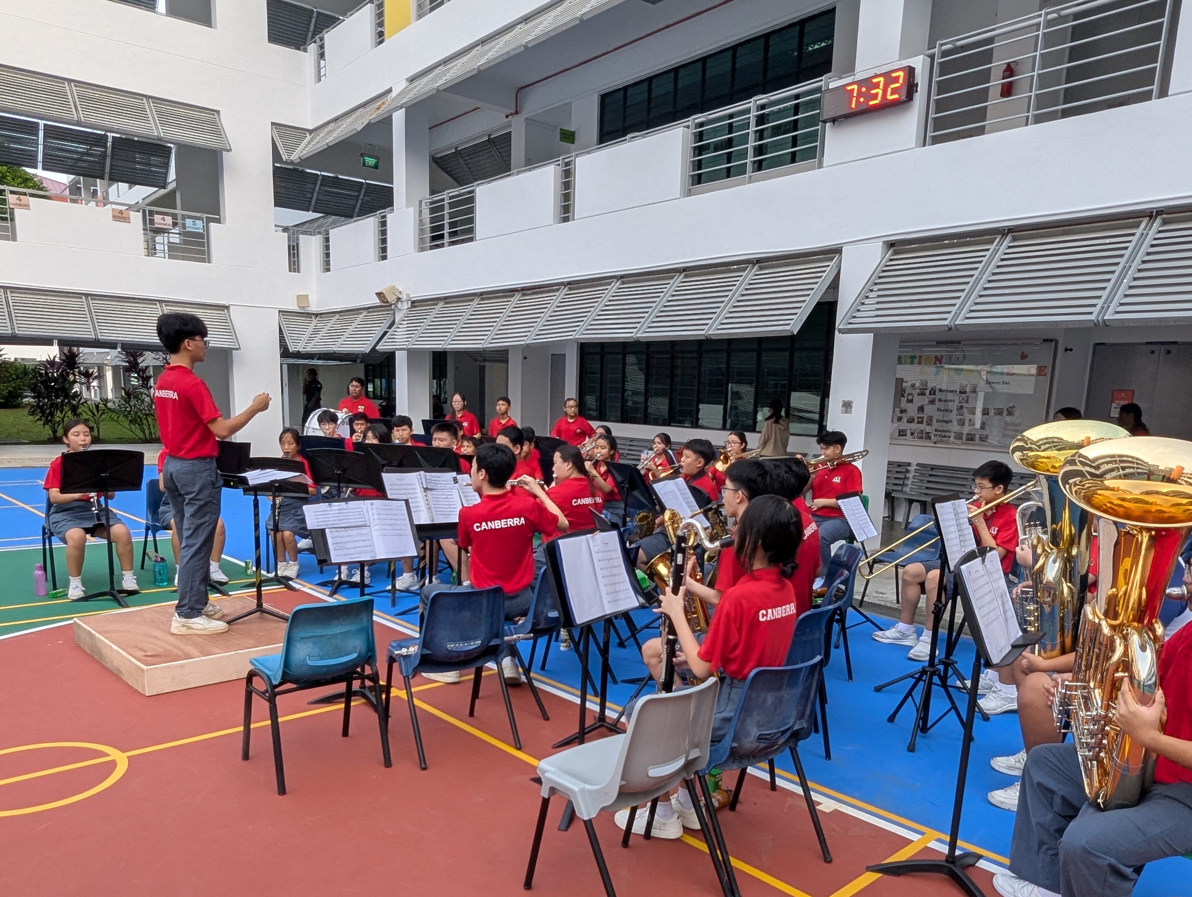 Students in red Canberra shirts play wind instruments outdoors on a school court, led by a conductor on a podium.
