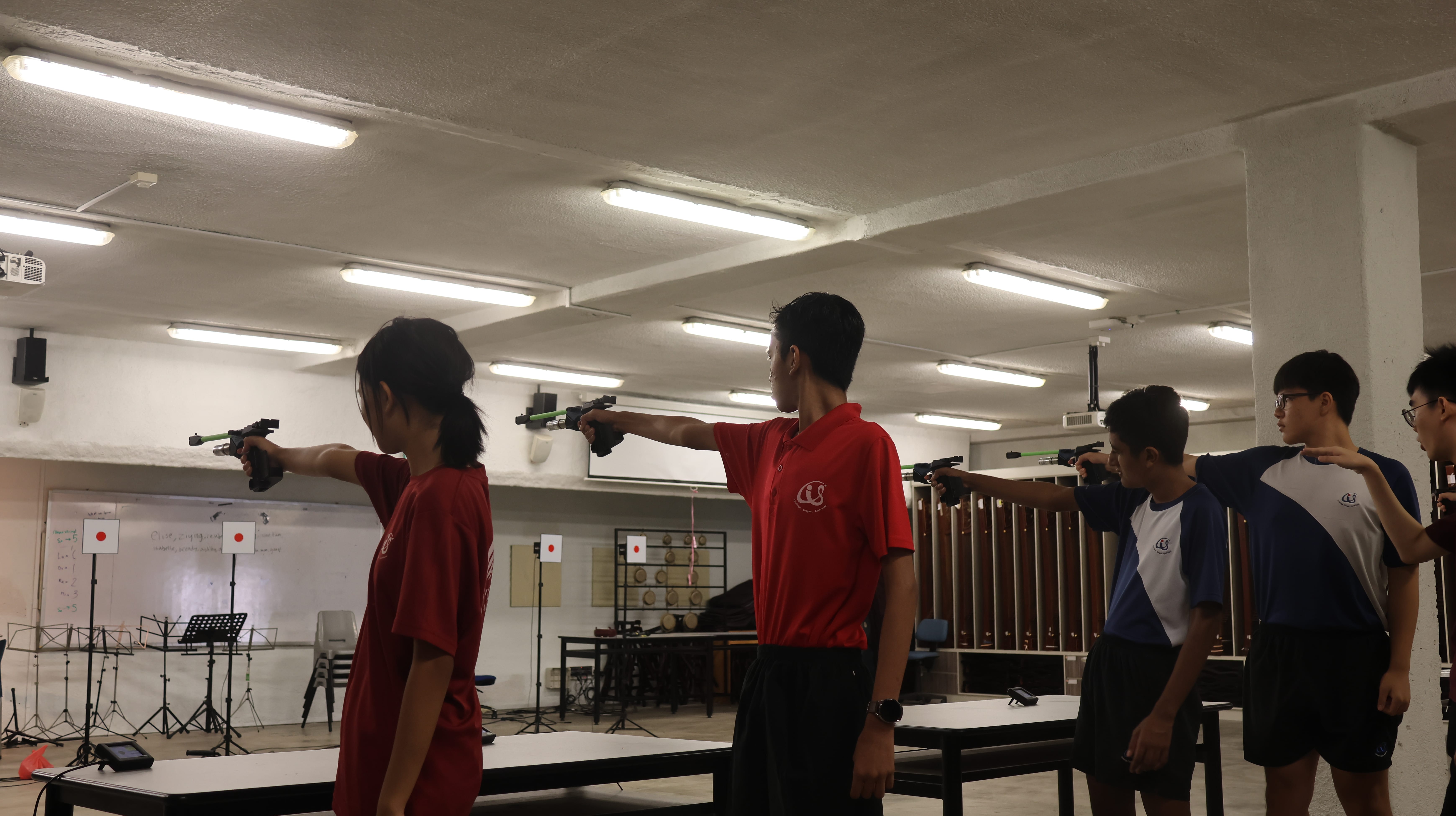 Several students in red and white uniforms aim pistols at paper targets in an indoor shooting range.