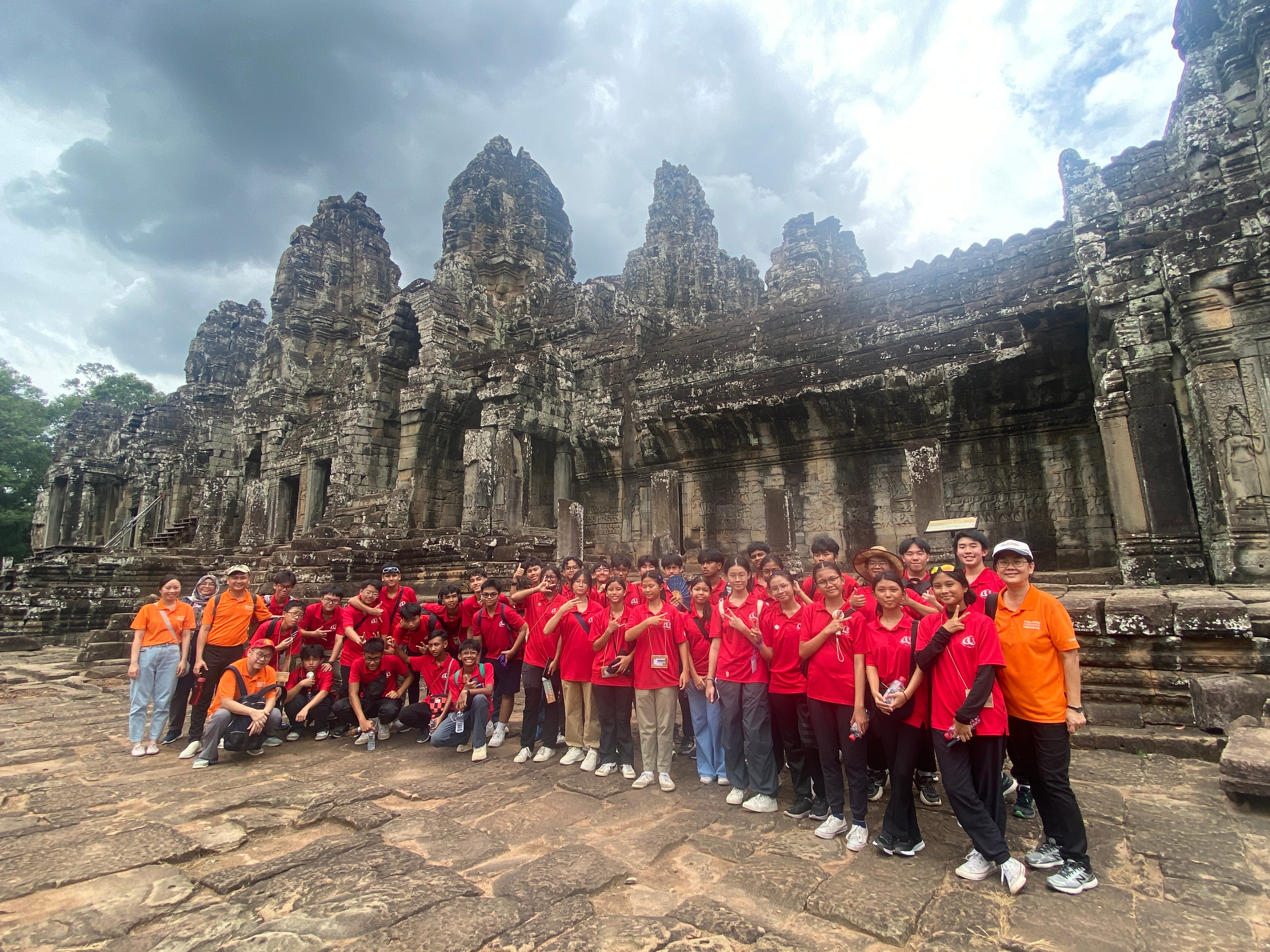 A large group in red and orange shirts poses in front of the ancient stone Bayon temple under a cloudy sky.