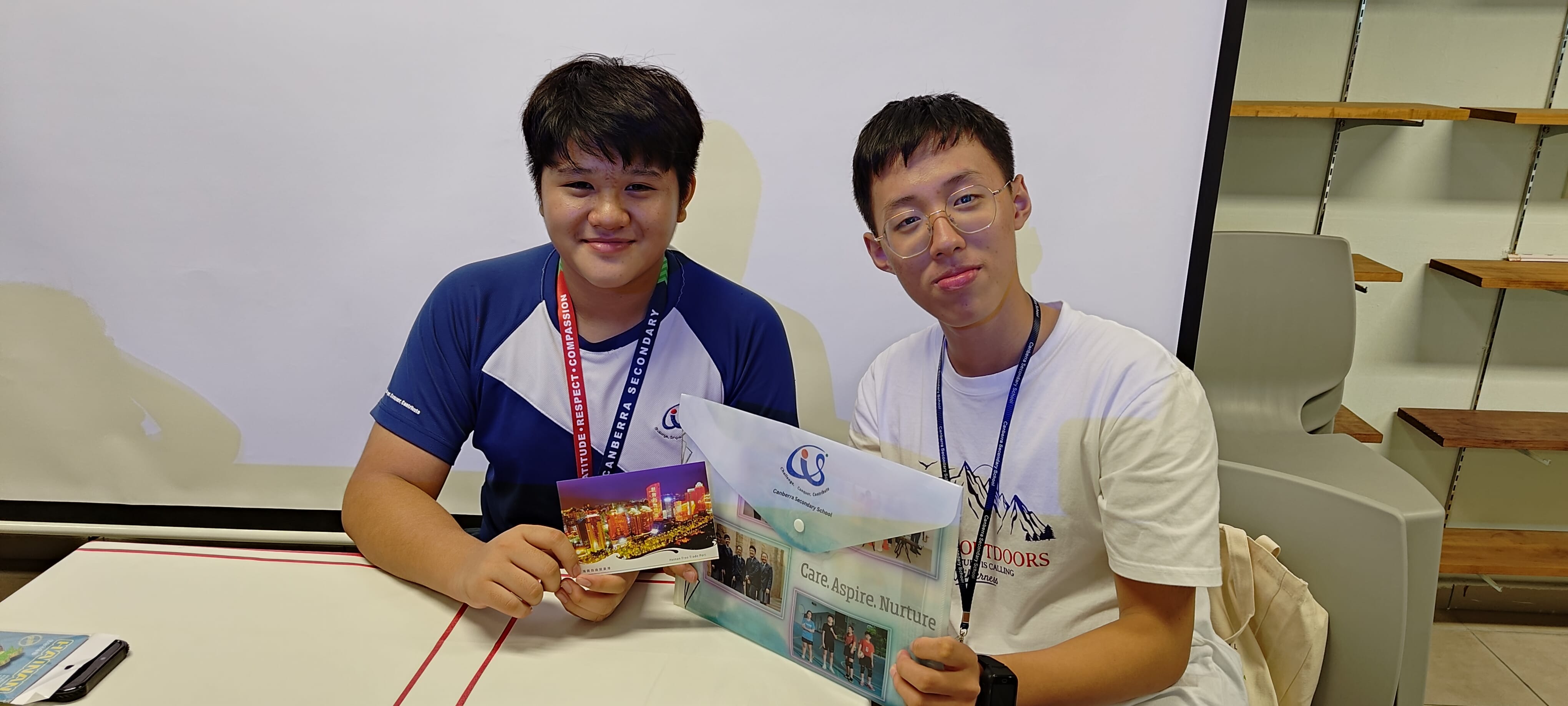 Two young male students wearing lanyards hold a Canberra Secondary School folder and a postcard while seated at a table.