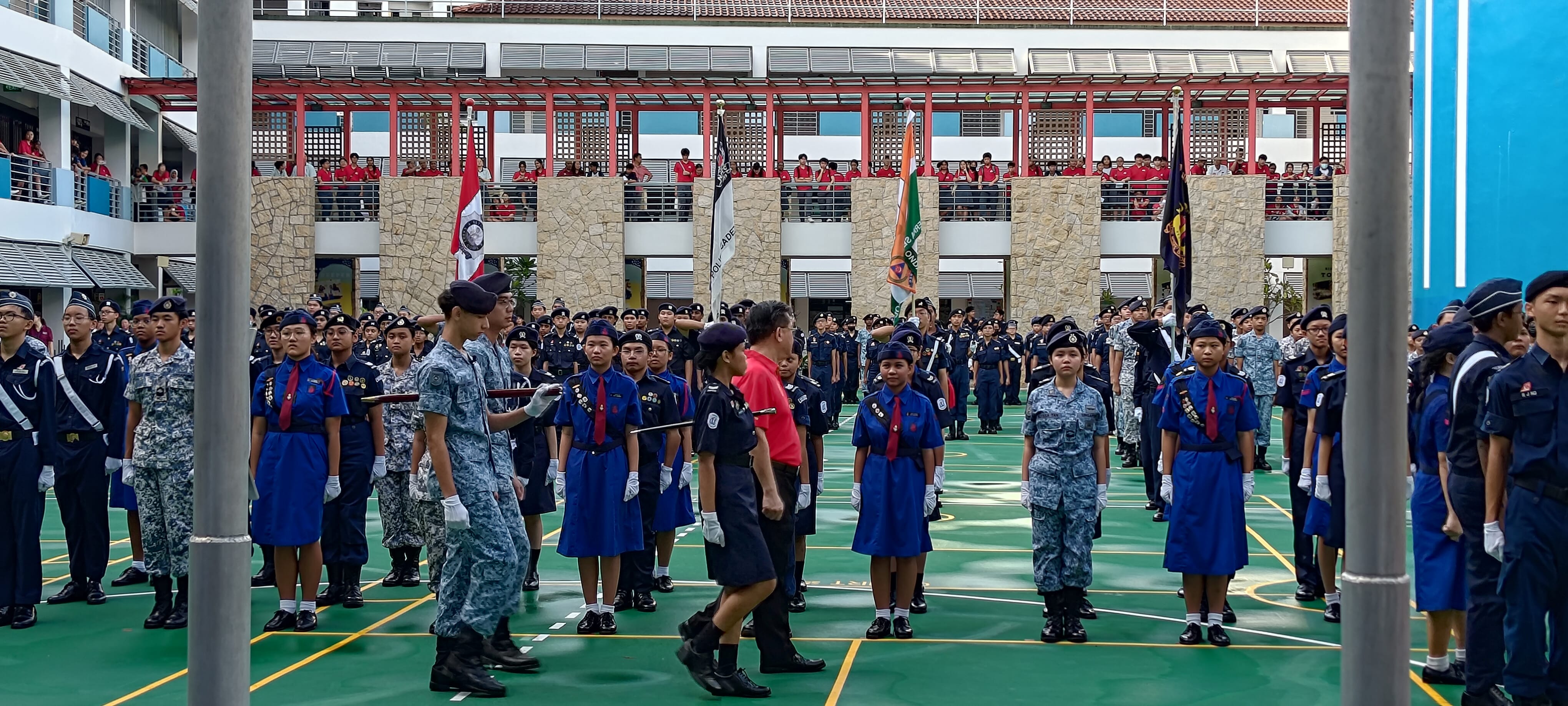 Students in various uniforms stand in formation during a school parade on a green court, with spectators on upper floors.