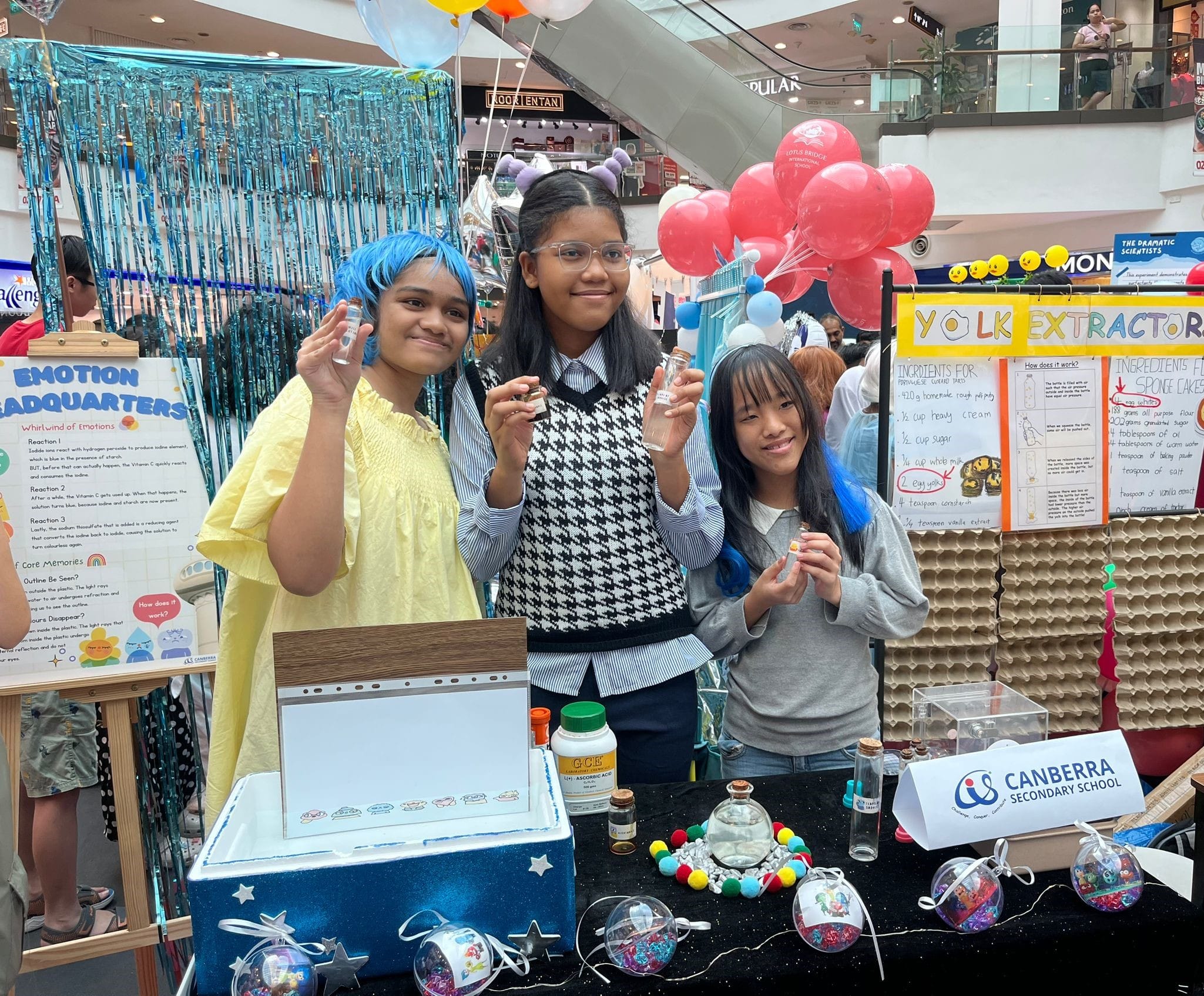 Three girls hold small vials at a Canberra Secondary School science fair booth in a mall.