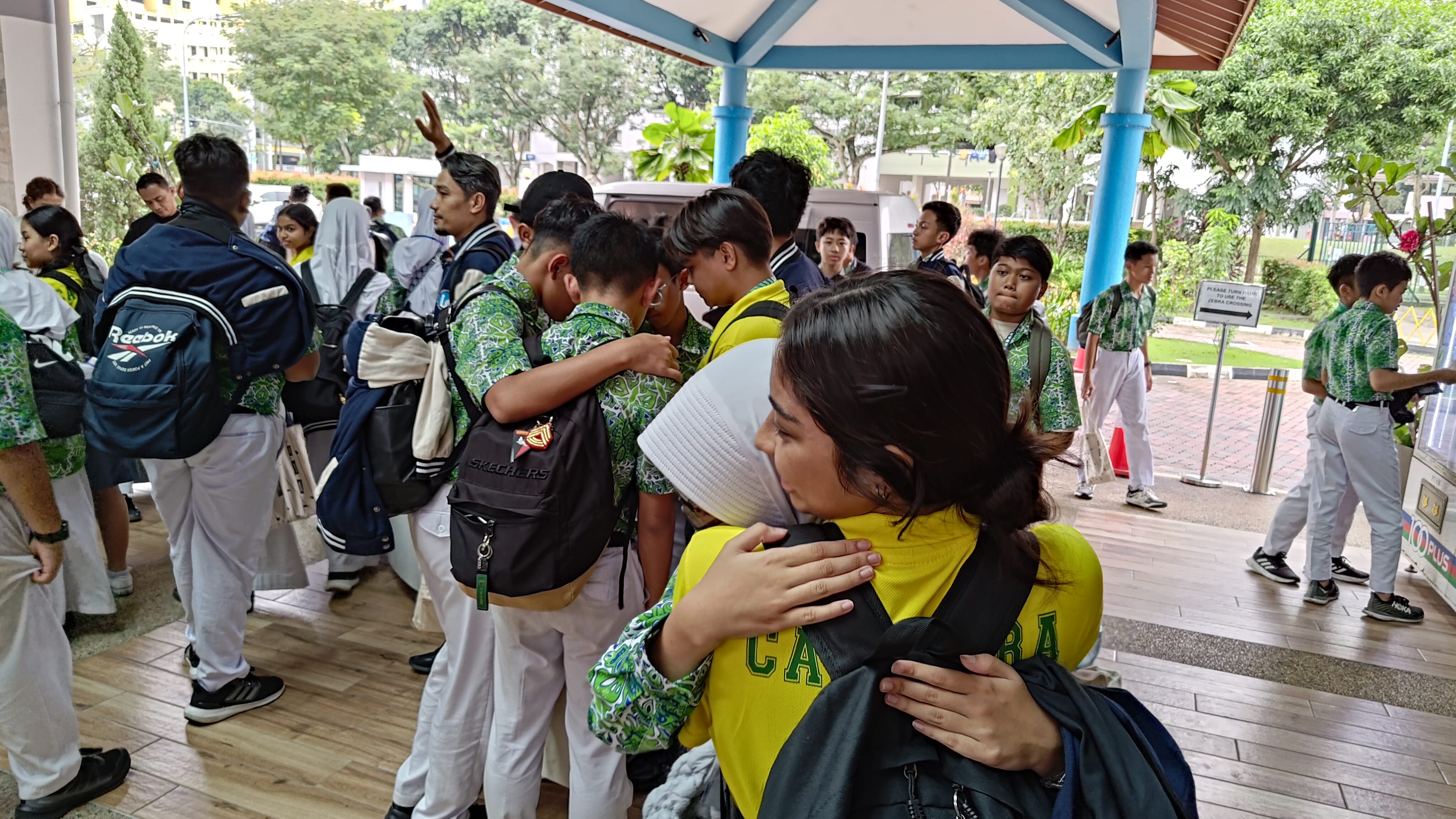 Students in green patterned uniforms hug and gather under a covered walkway outdoors near trees and buildings.