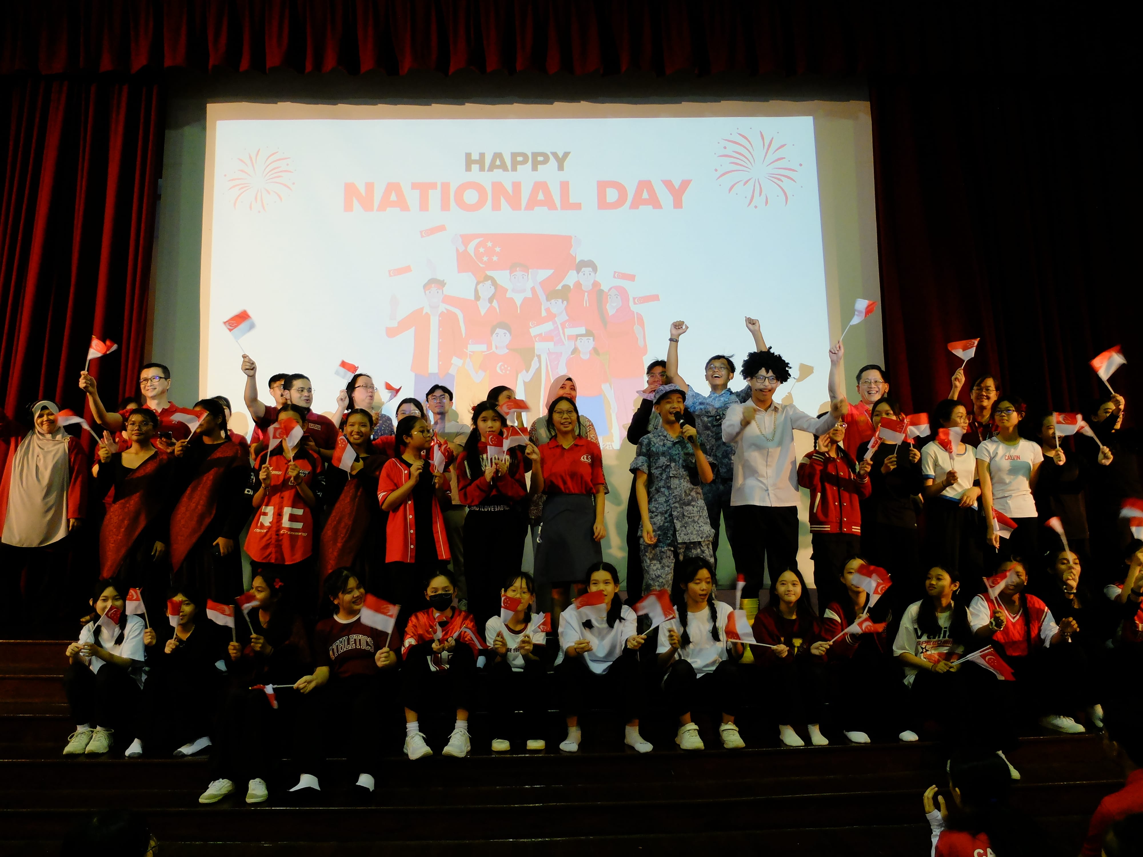 Large group on stage waving Singapore flags in front of a screen reading Happy National Day.