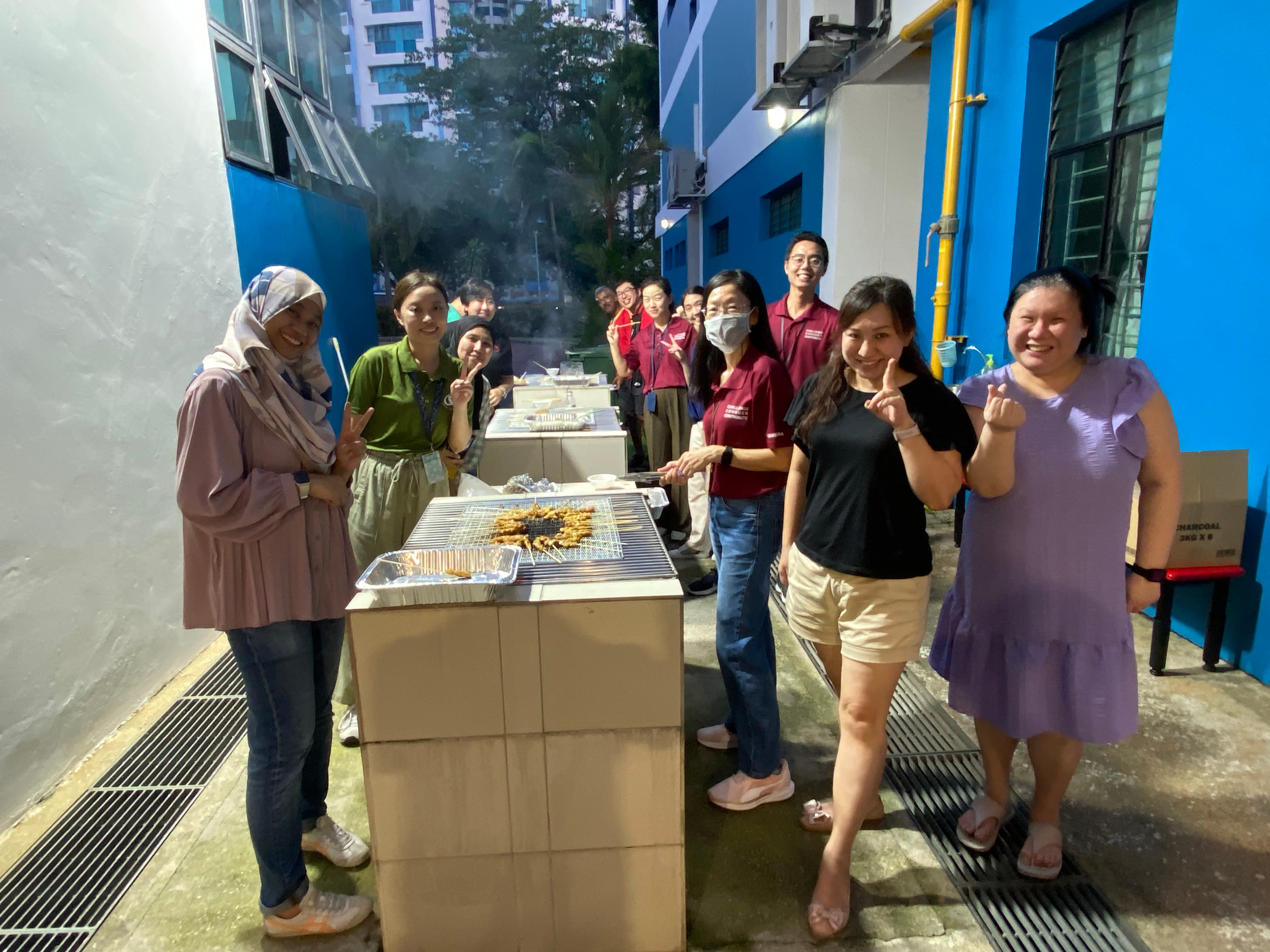A group of people smiling and posing at an outdoor barbecue grill at night near blue buildings.