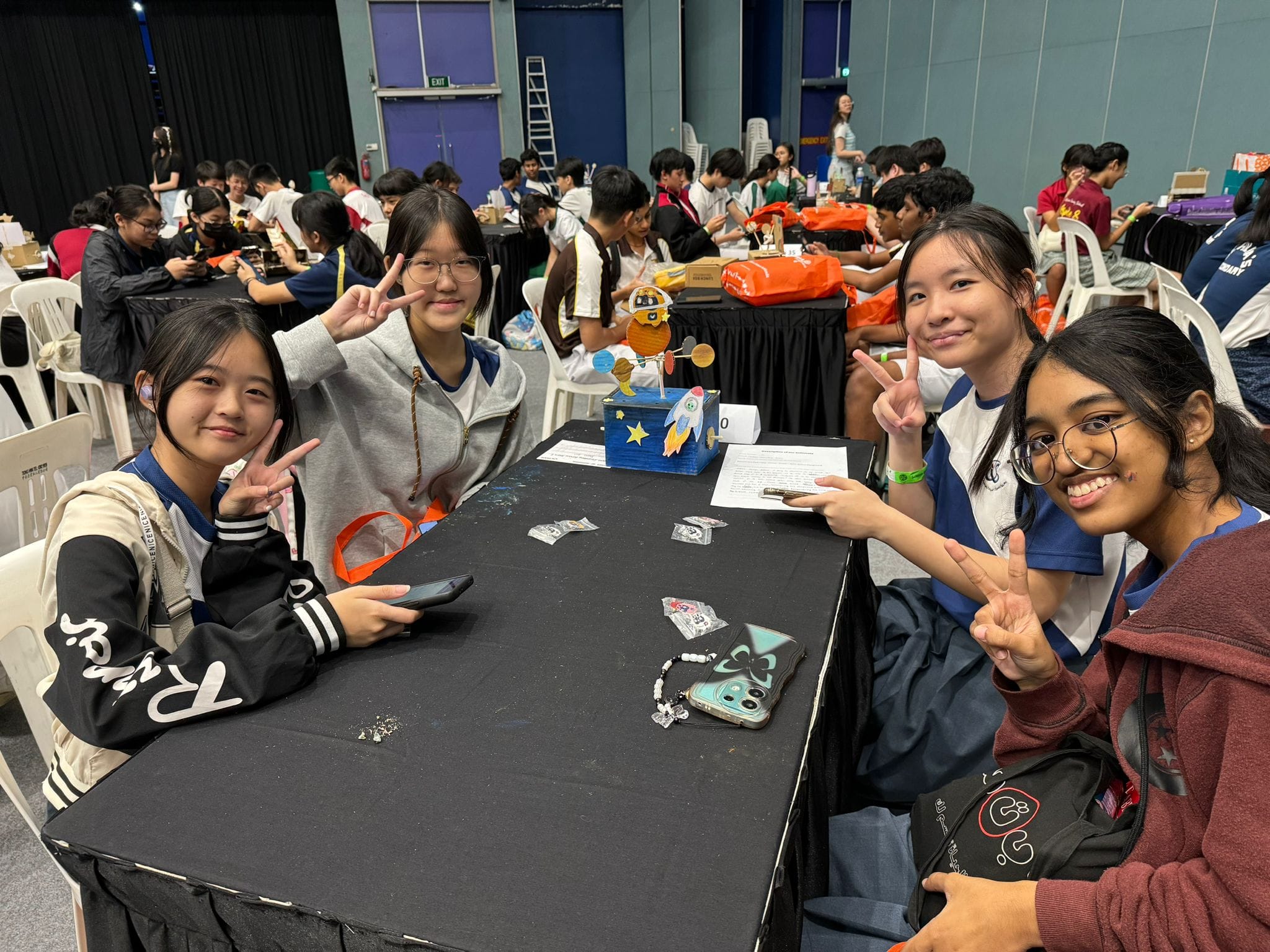 Four teenage girls smile and pose with peace signs at a table displaying a space-themed craft project in a busy hall.