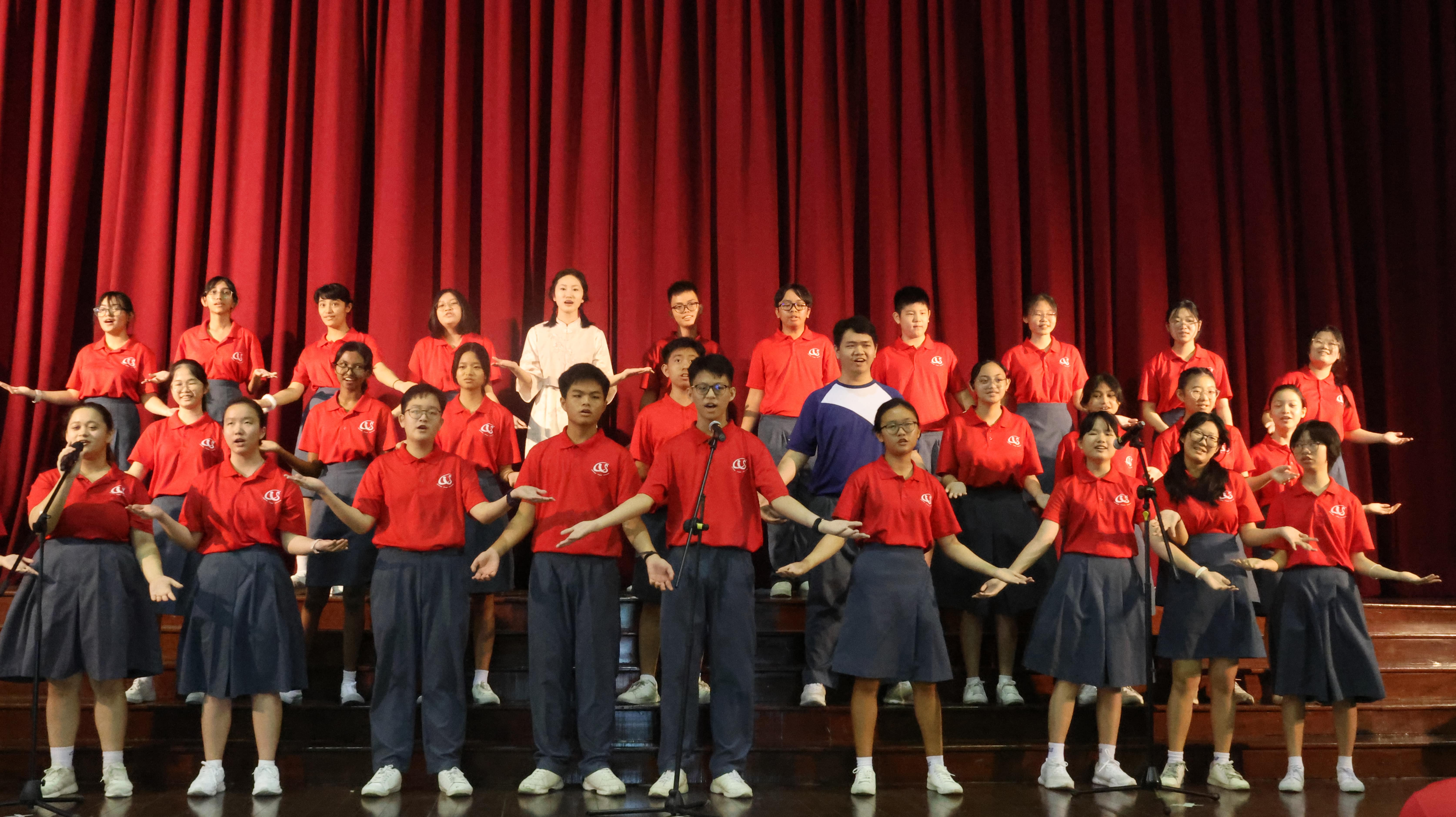 A large student choir in red uniforms performs on stage with arms outstretched before a red curtain backdrop.