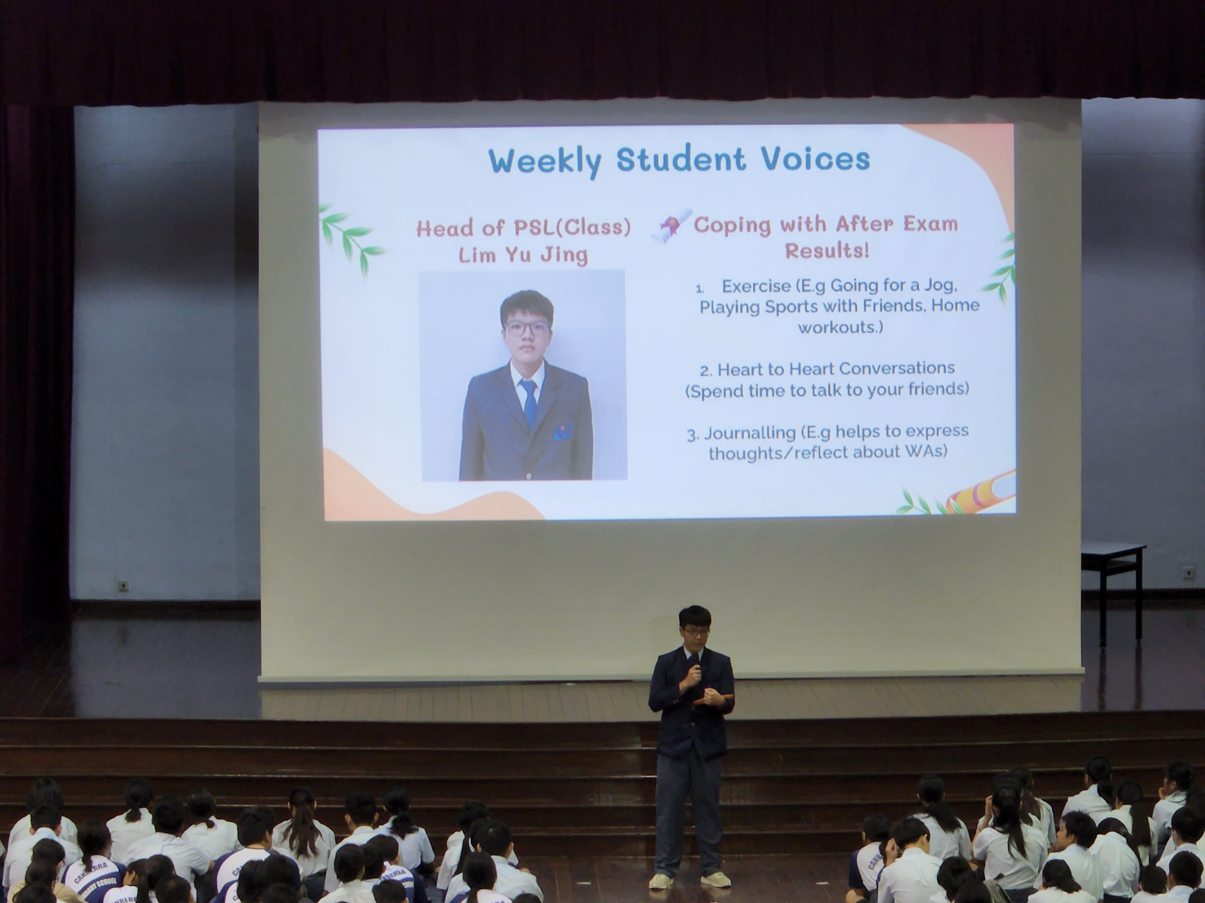 Boy in school uniform and blazer addressing the school, with an image of him as well as talking points on a screen behind him