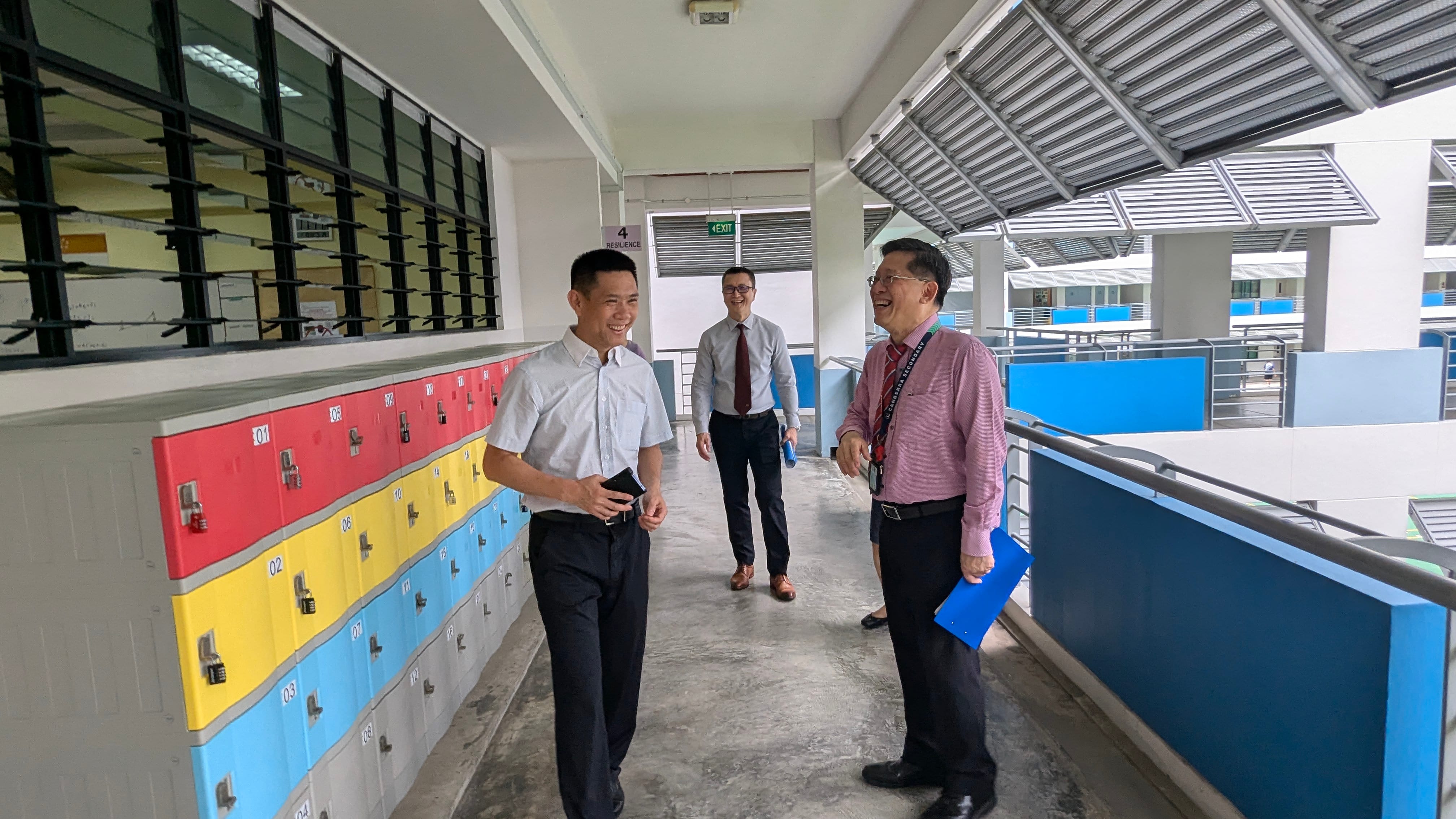 Three men in formal shirts walk and laugh in a school corridor beside colorful red, yellow, and blue lockers.