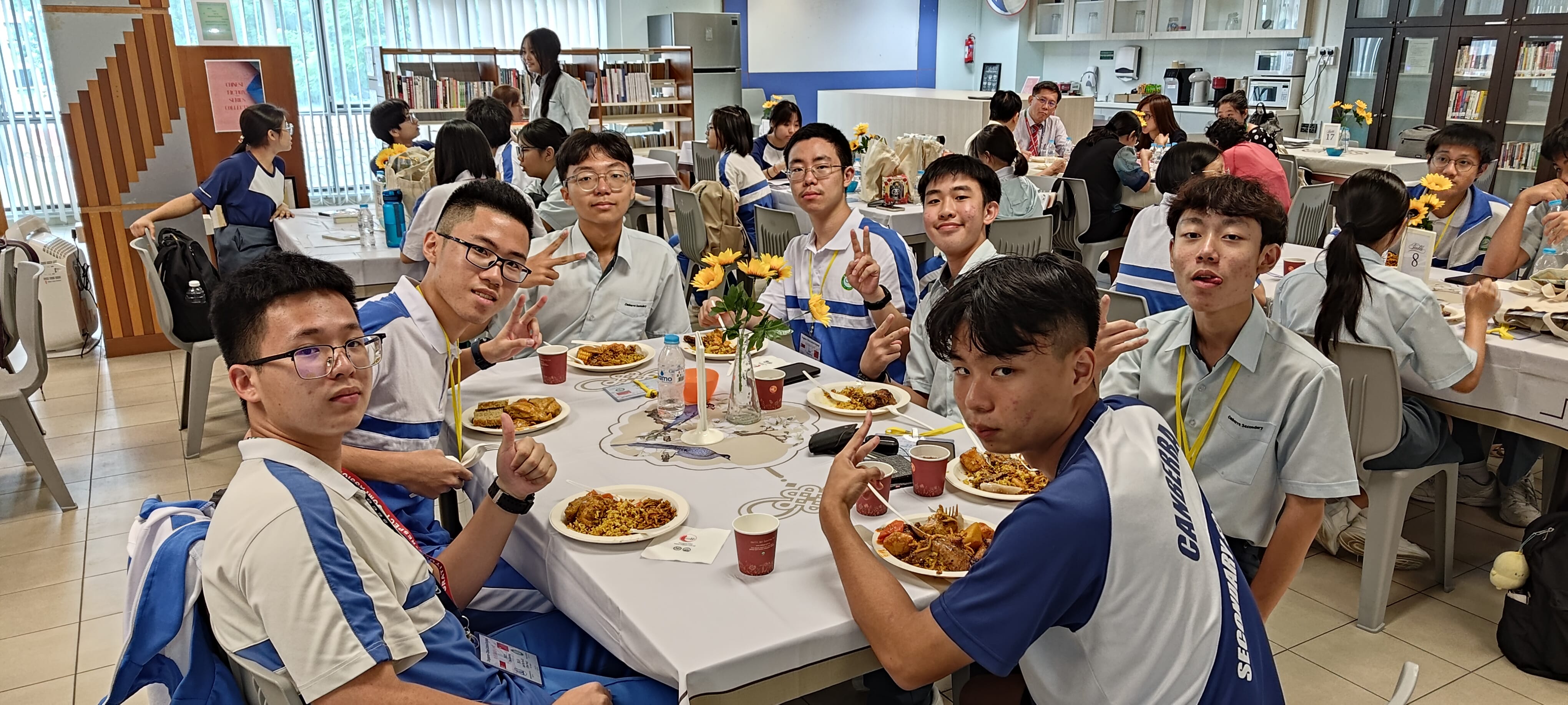 Students in blue and white uniforms sit together at a table with food, posing with peace signs in a school cafeteria.