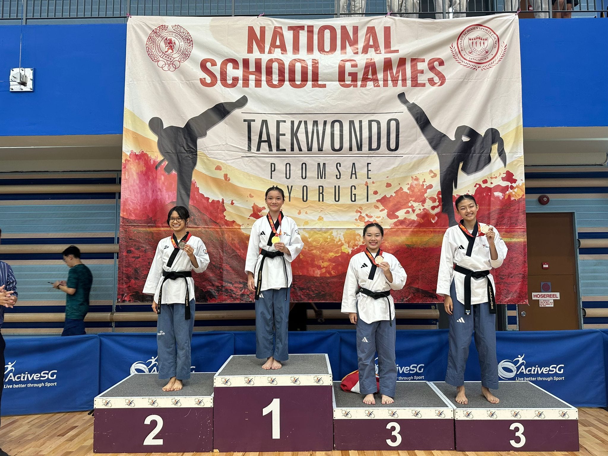 Four taekwondo athletes in white uniforms stand on a podium at the National School Games Taekwondo event, holding medals.
