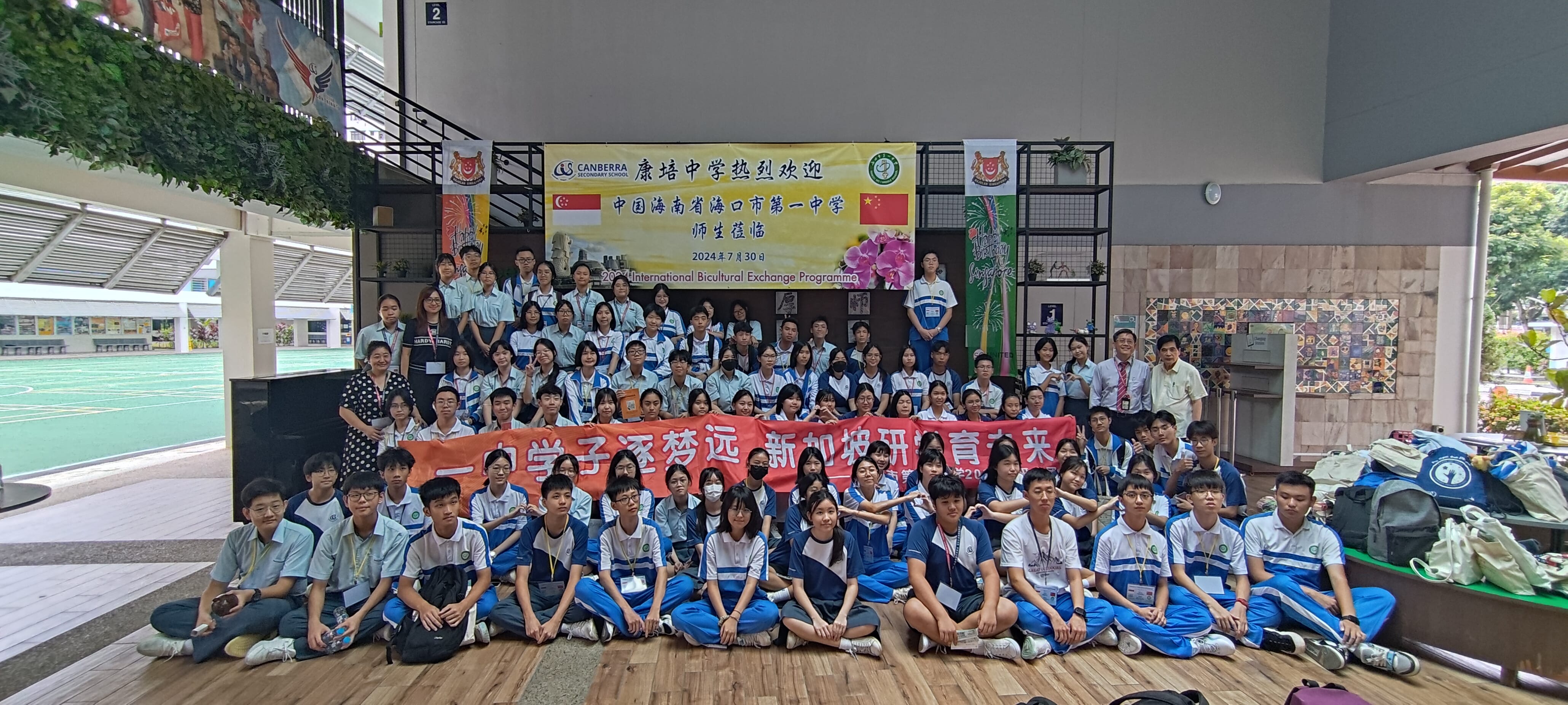 Large group of students in blue uniforms posing with a red Chinese banner at a 2024 bicultural exchange programme event.