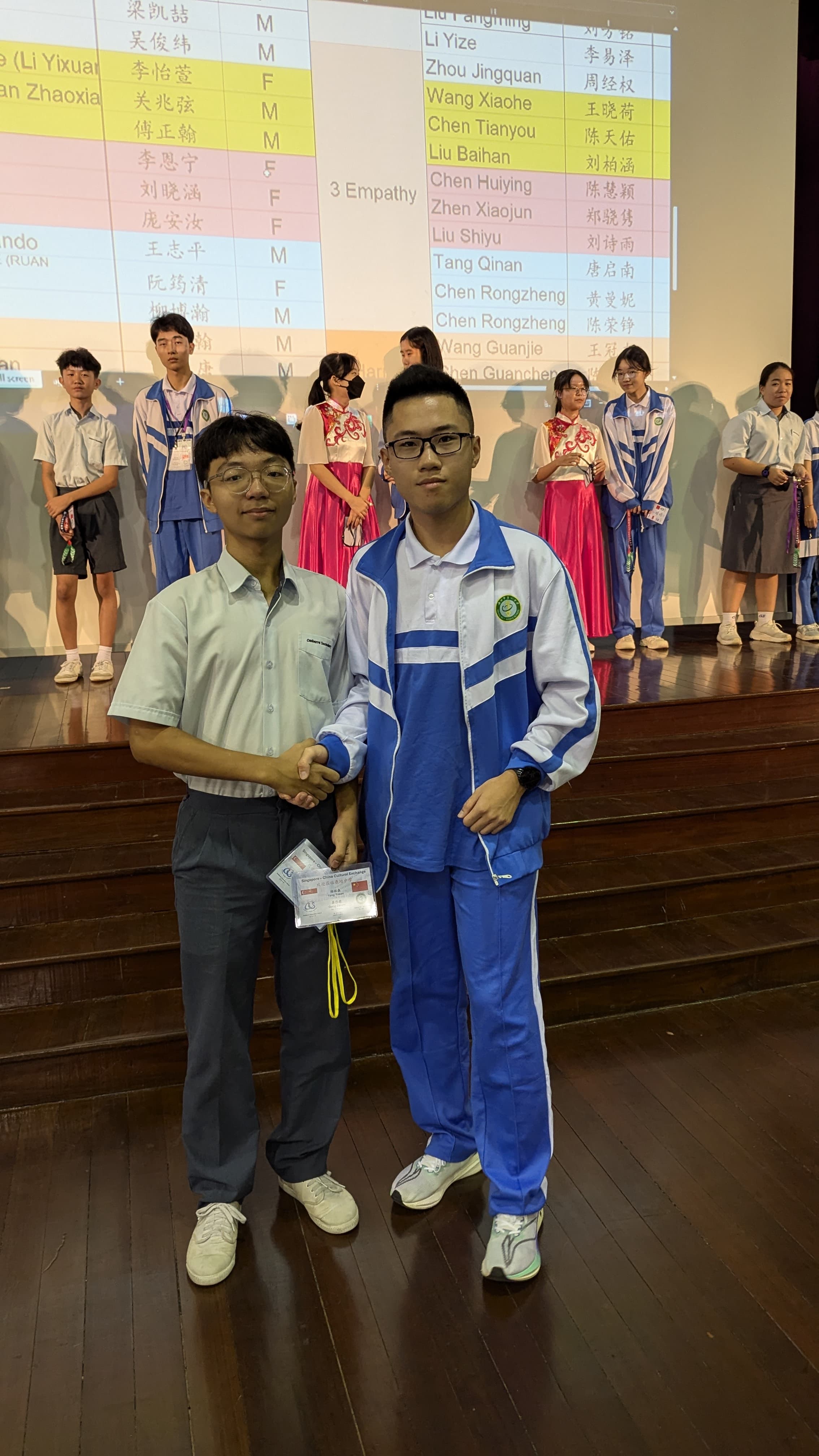 Two young men shaking hands on a stage with a projected name list and students in uniforms behind them.