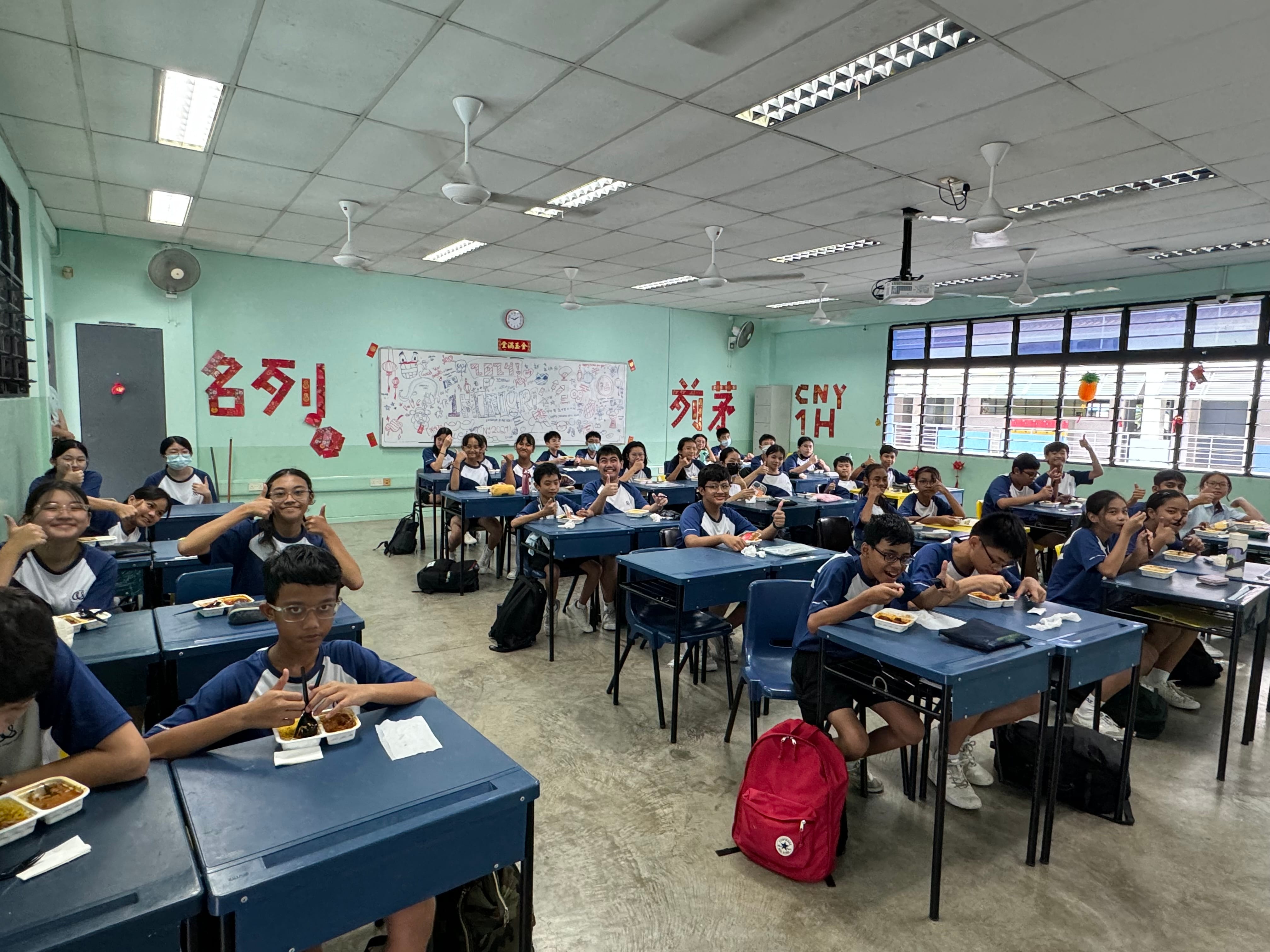 Students in blue uniforms sit at desks eating food in a decorated classroom, smiling and giving thumbs up.