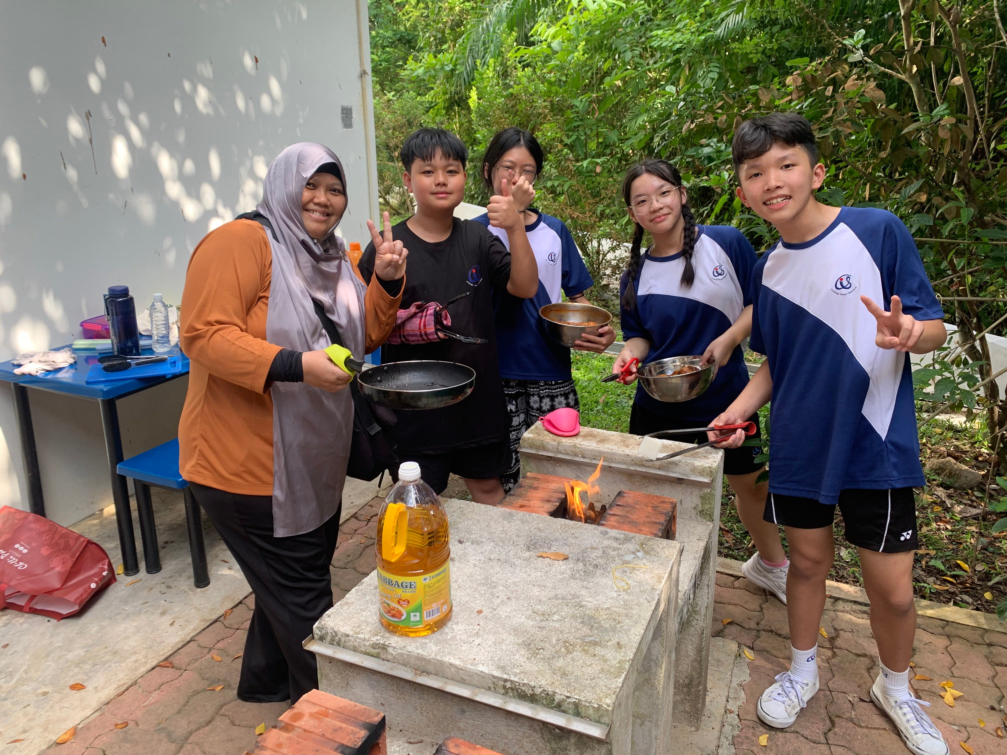 Five people smile and pose while cooking over an open-fire outdoor stove, holding pans and bowls outdoors.
