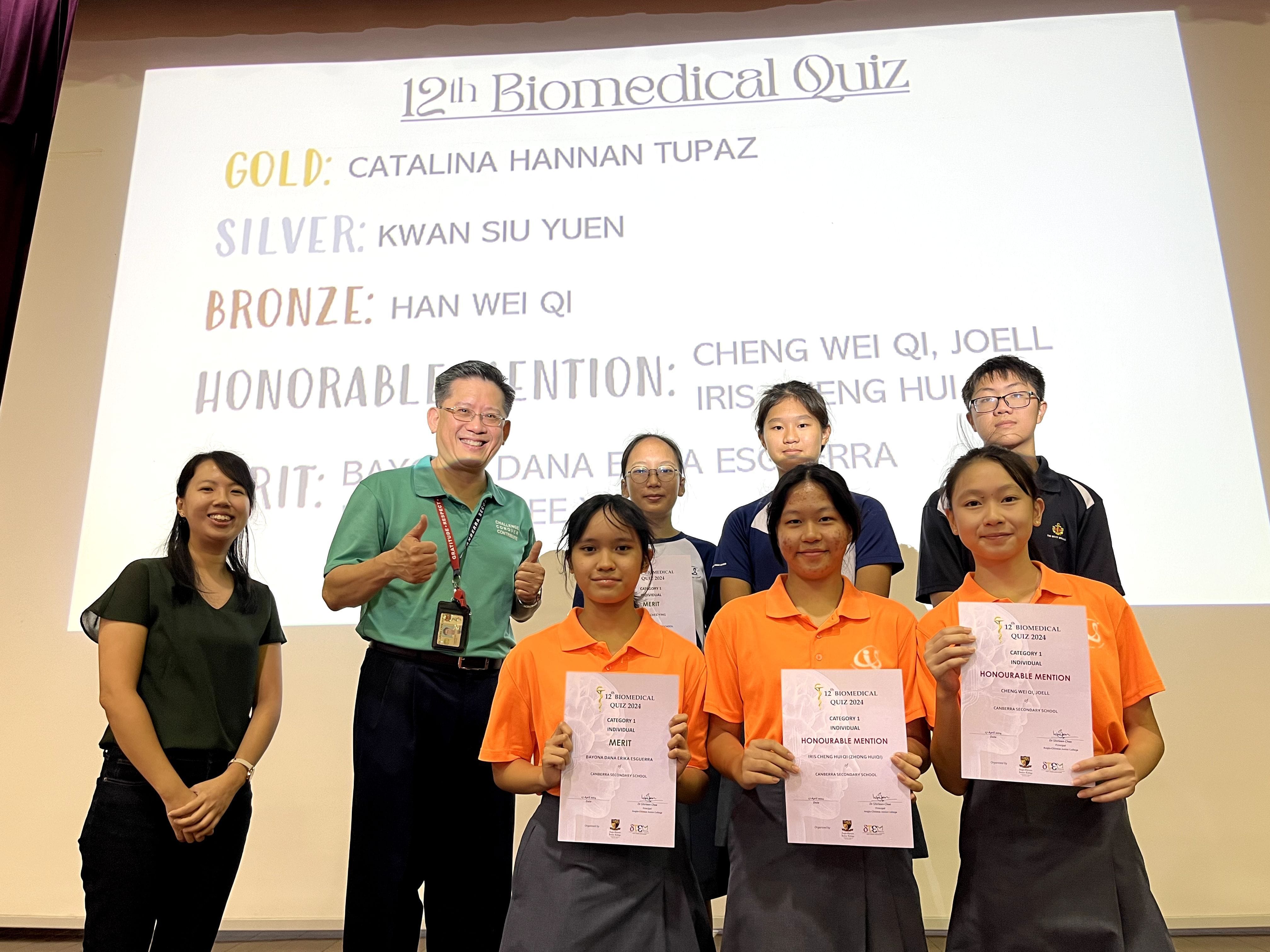 Students and teachers pose with certificates in front of a 12th Biomedical Quiz winners slide.
