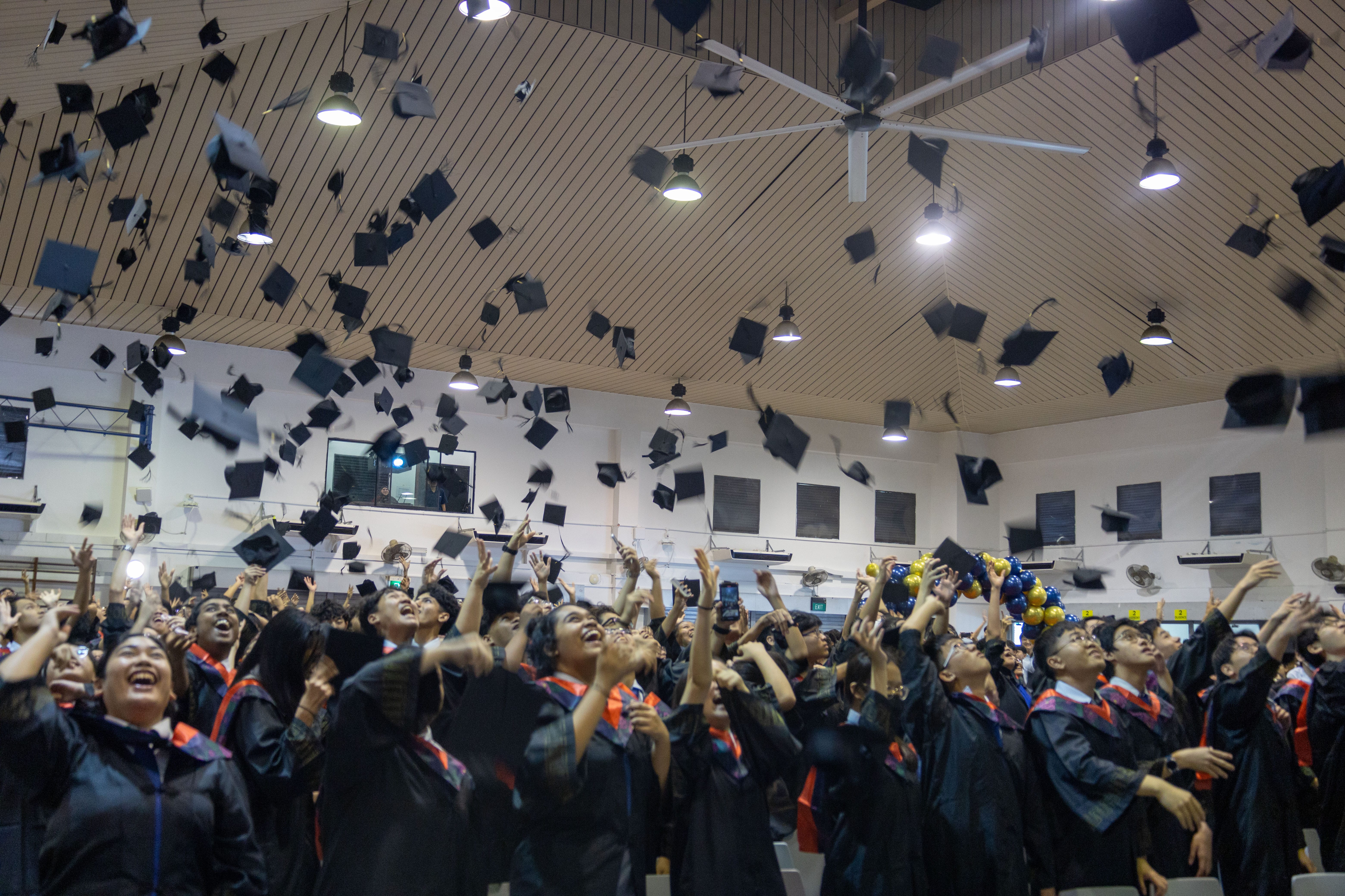 Graduates in gowns cheer as many mortarboards fly overhead inside a hall with bright ceiling lights.