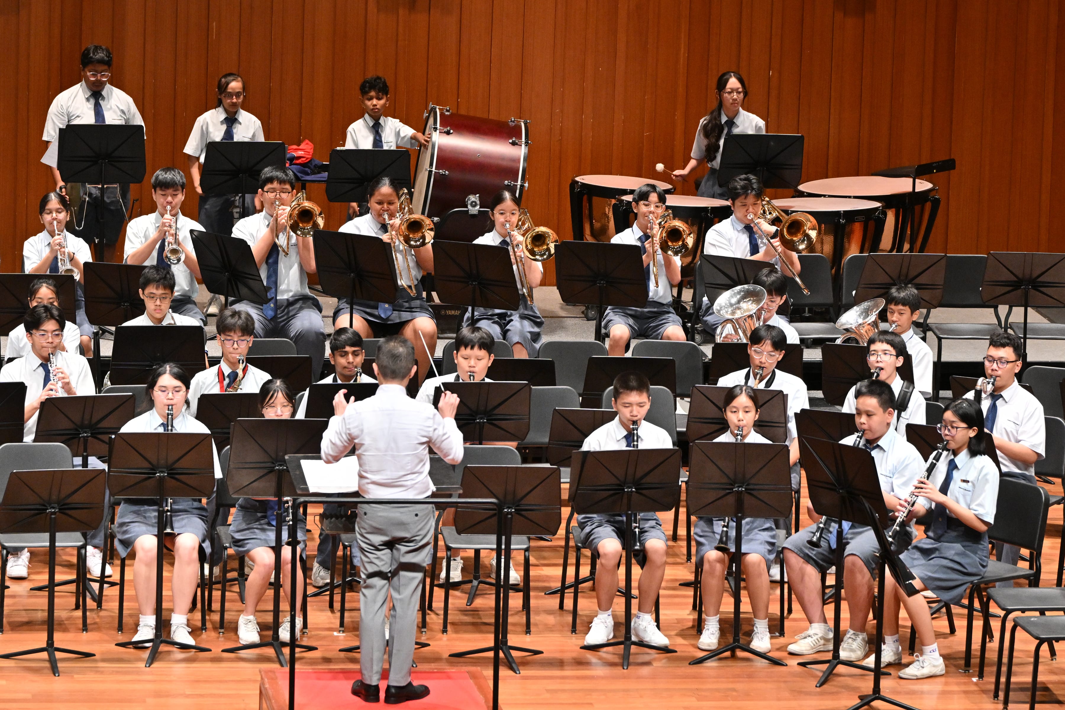 Conductor leads a school band in uniforms with brass, woodwinds, and percussion on a stage.