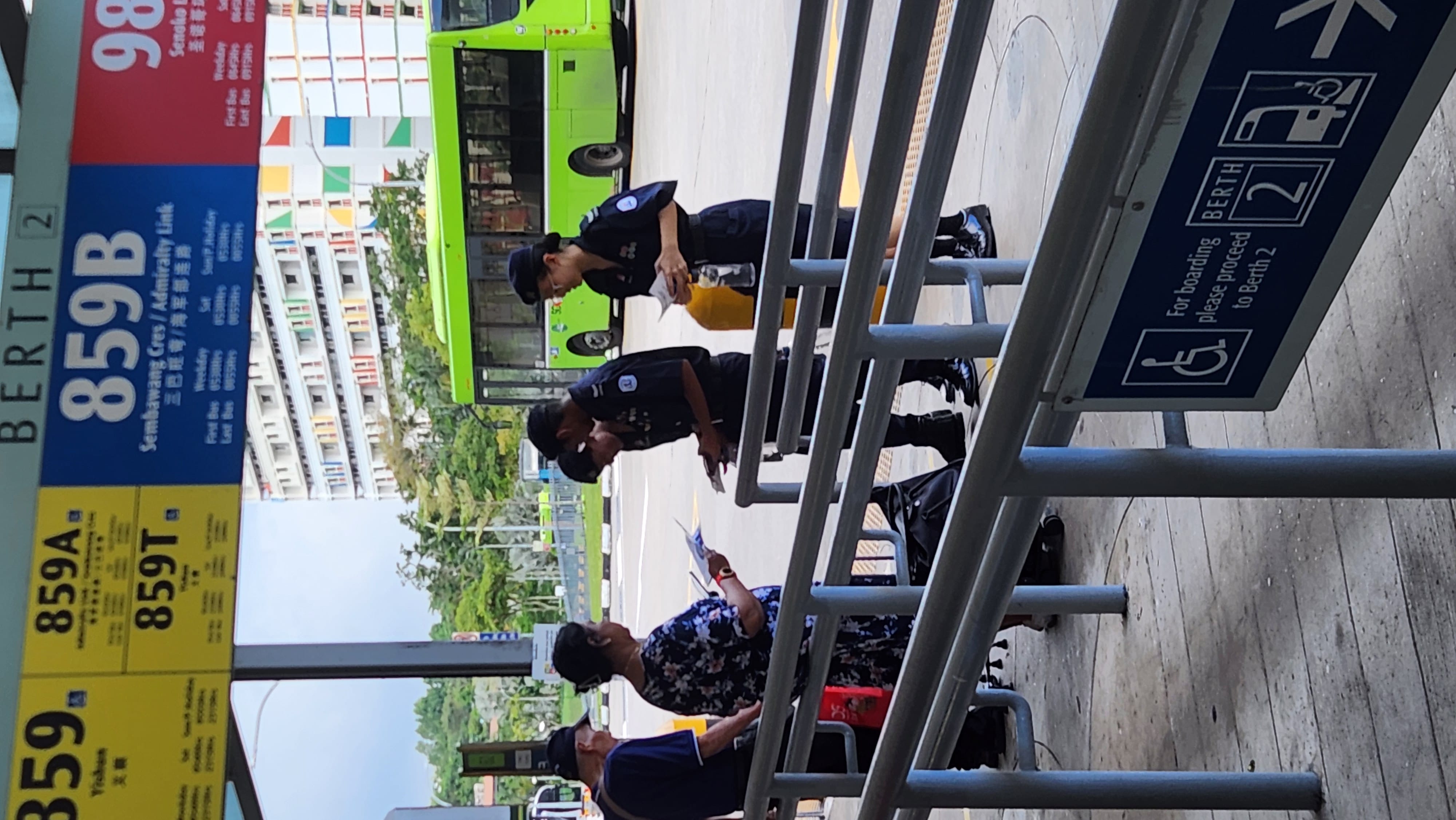 Two uniformed officers hand flyers to two civilians at a Singapore bus interchange, Berth 2, with a green bus behind.