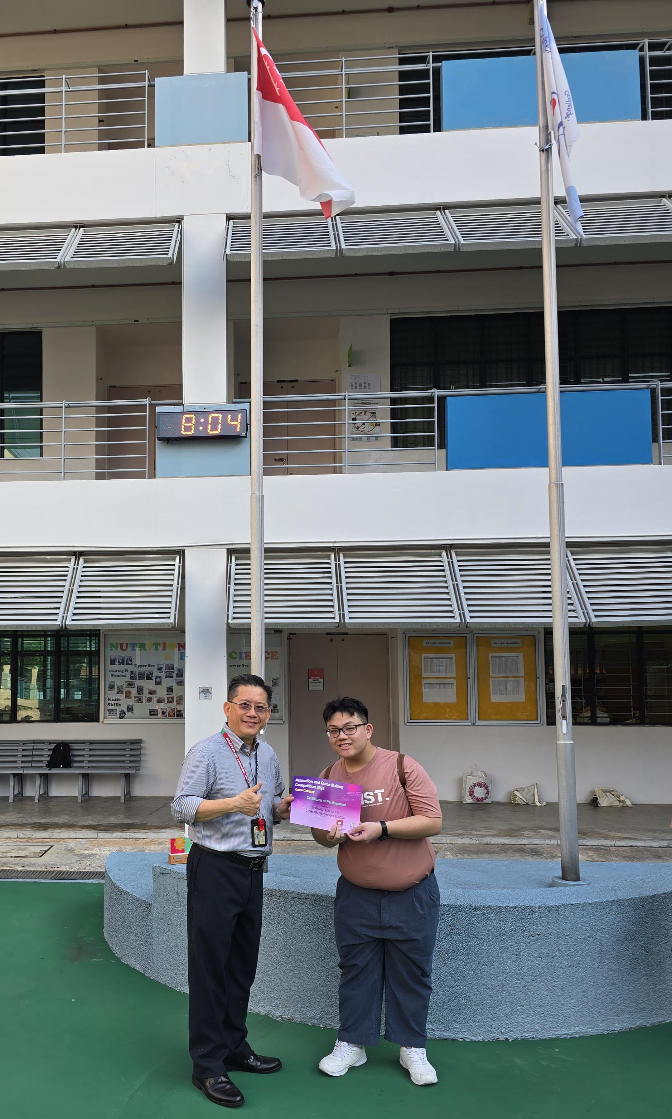 Two people stand in a school courtyard holding a certificate near flagpoles with Singapore and school flags.
