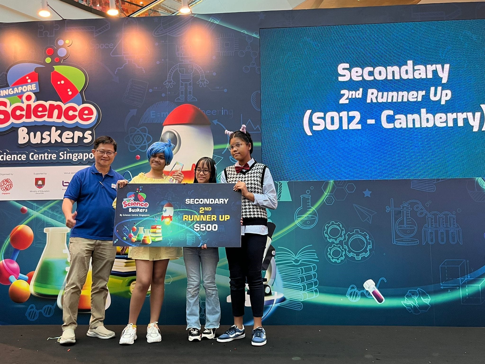 Three people pose on stage holding a Singapore Science Buskers Secondary 2nd Runner Up $500 prize board.