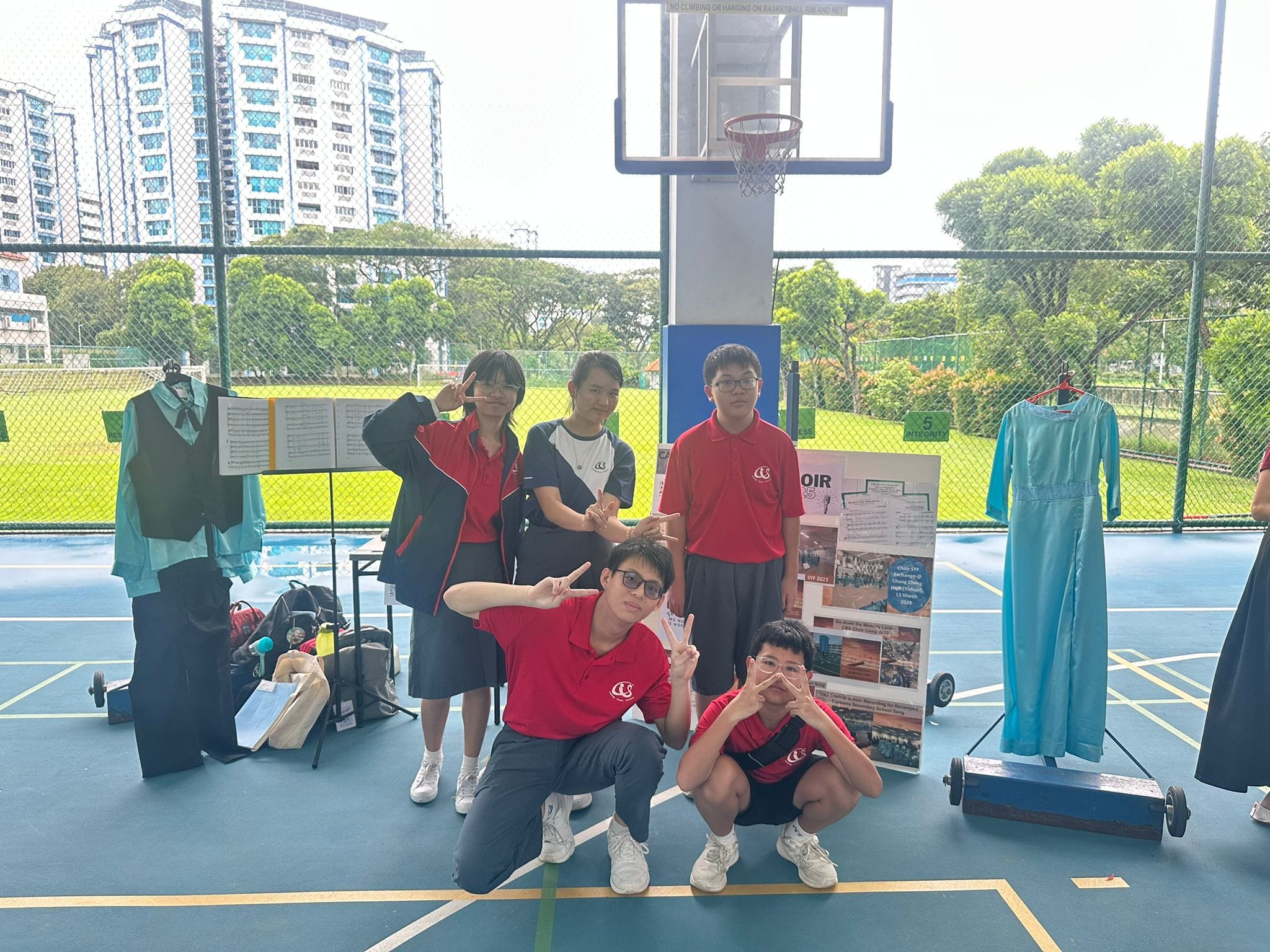 Five students in red uniforms pose on an outdoor basketball court beside a choir display with costumes and sheet music.