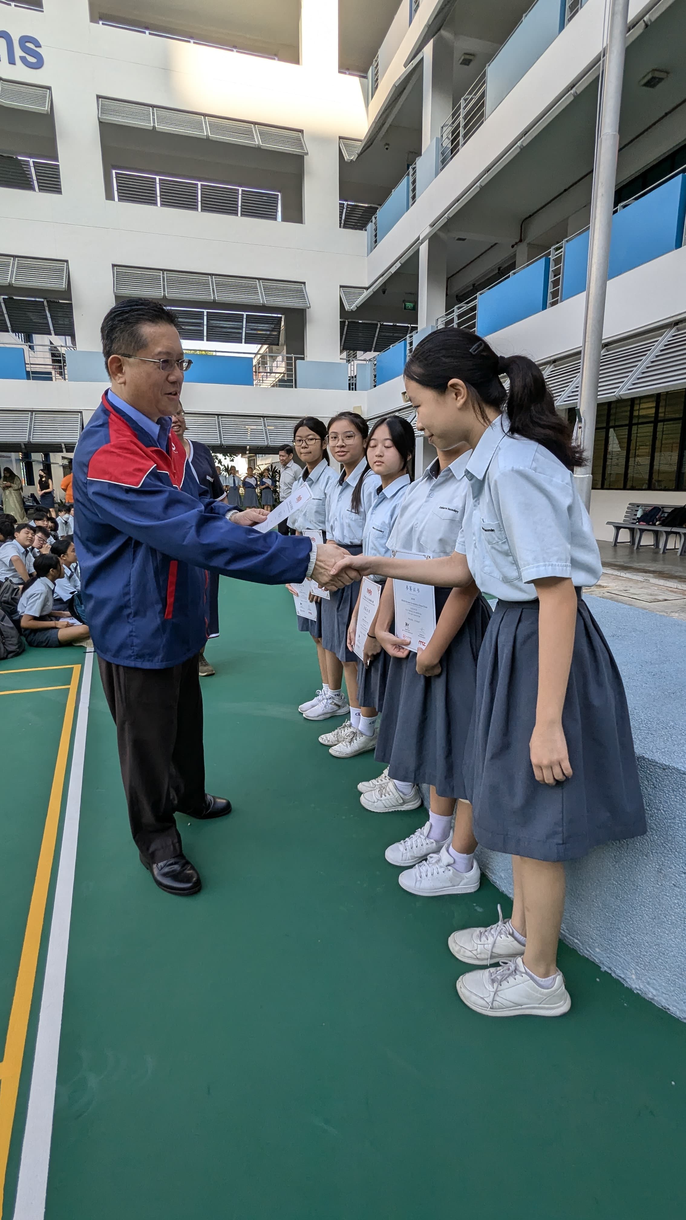 A man in a blue jacket shakes hands with a female student while presenting certificates to a line of uniformed students outdoors.