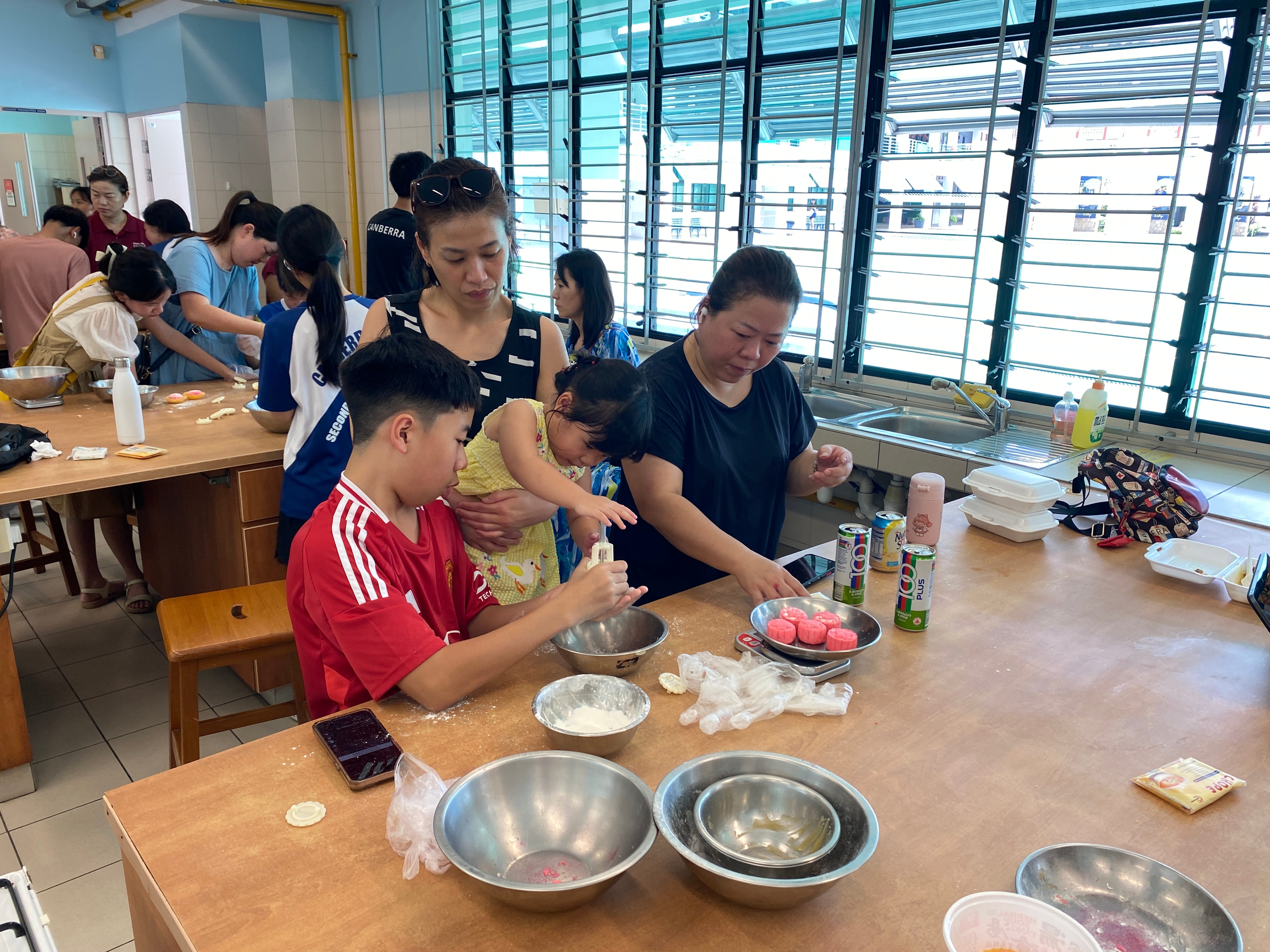 Adults and children gather around wooden tables making pink dumplings in a busy community kitchen.