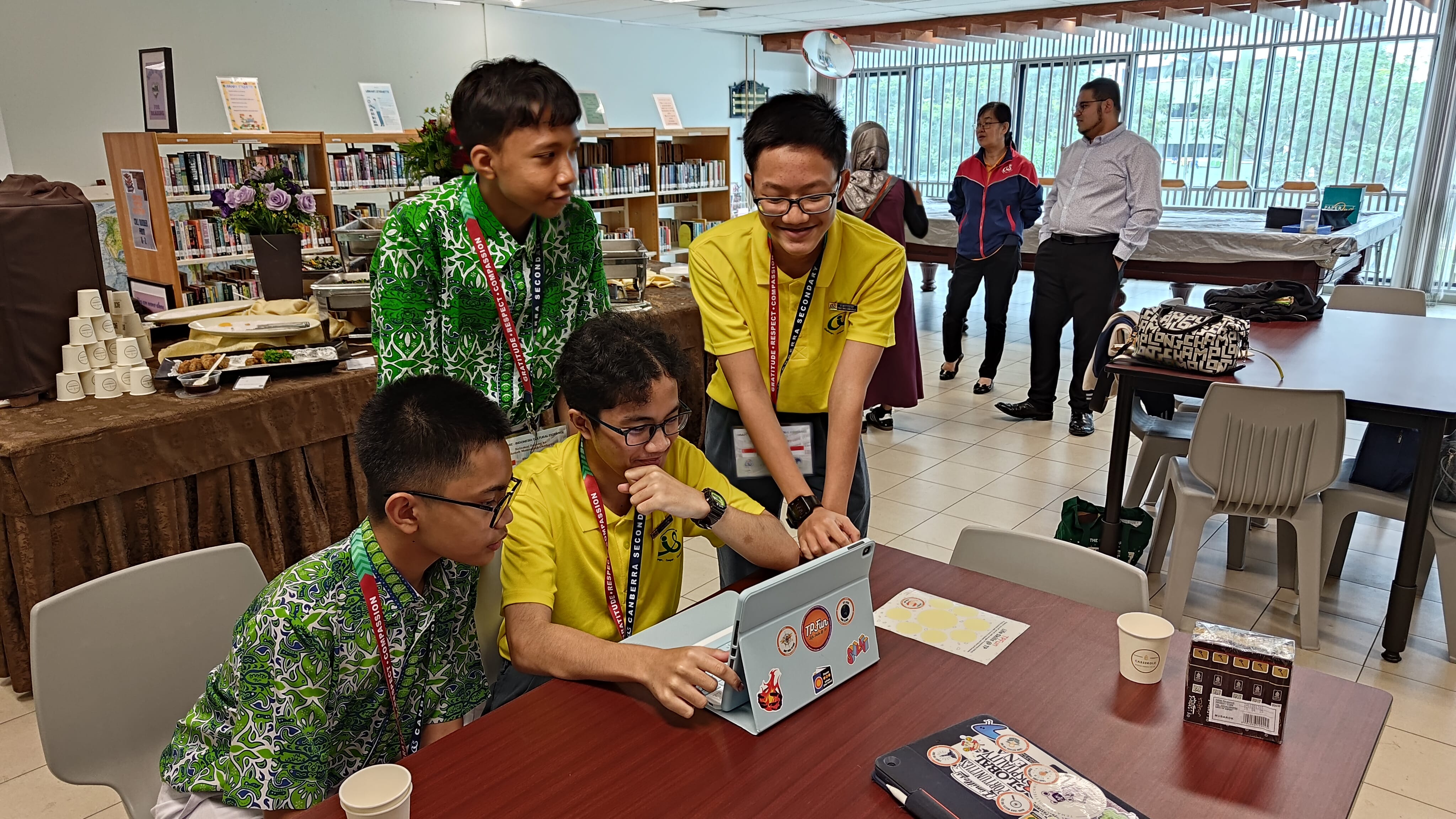 Four students huddle around a tablet at a table in a library, with adults standing in the background.
