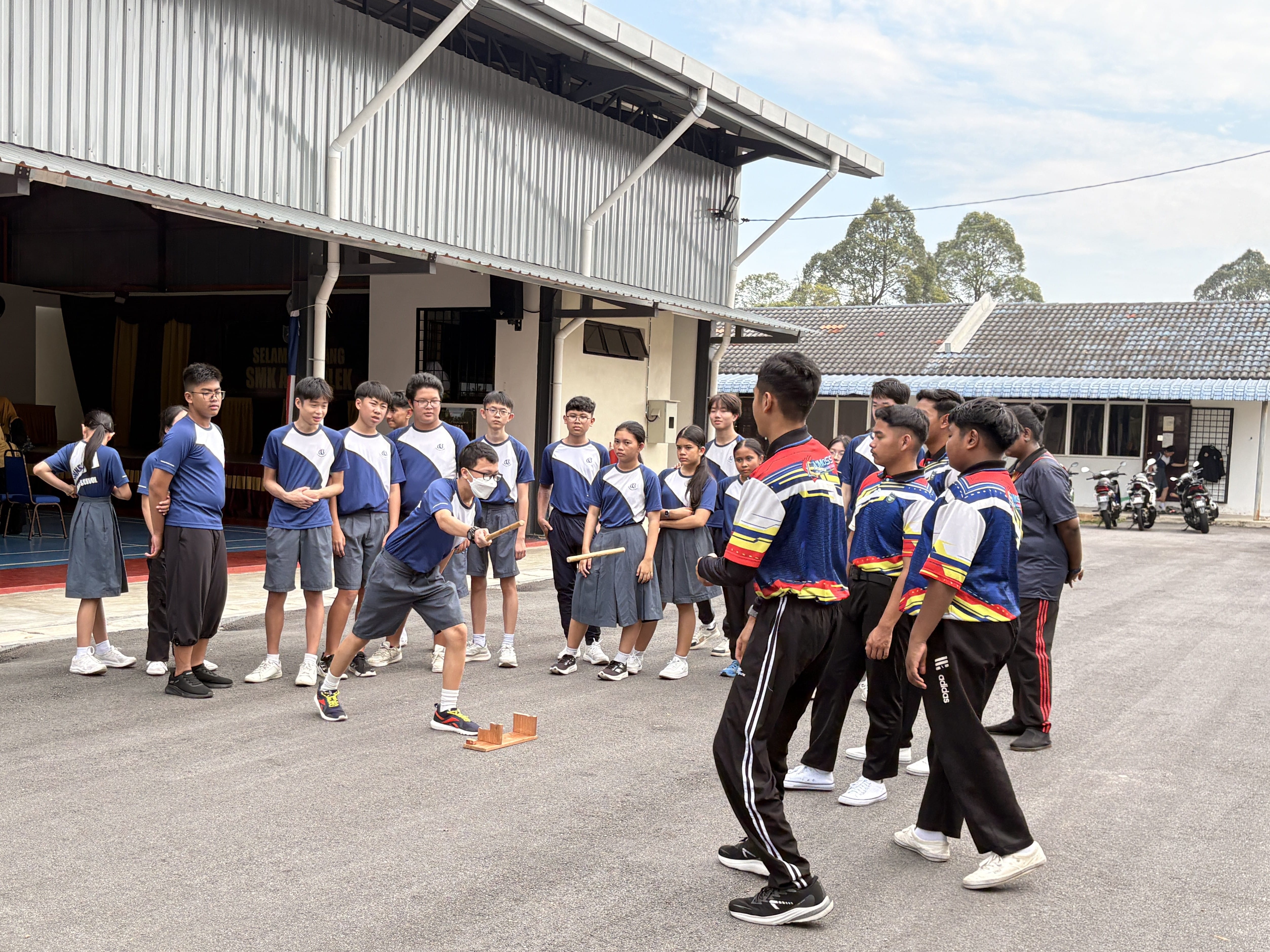 Students in blue uniforms watch a person demonstrate a traditional stick game on a school courtyard outdoors.