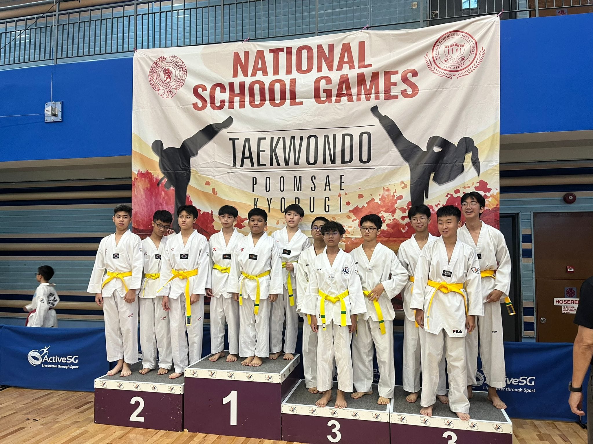 Young taekwondo athletes in white uniforms with yellow belts stand on a podium at the National School Games.