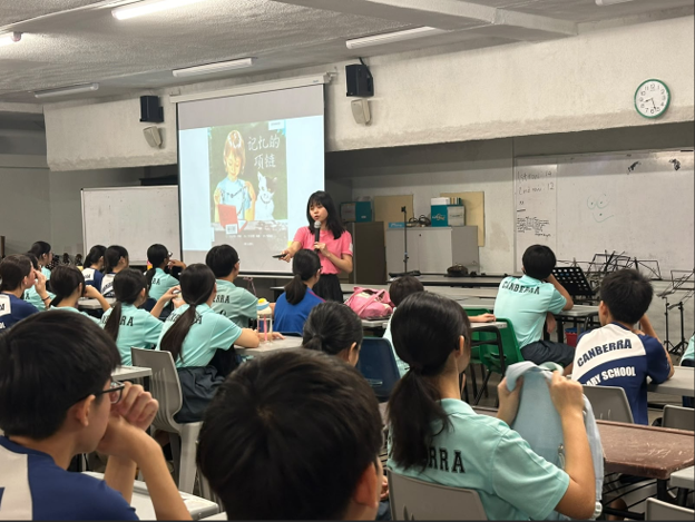 A teacher in a pink t-shirt stands at the front of a large classroom, holding a microphone and presenting to students. A projector screen behind her displays a Chinese book cover titled 《忘了的项链》. Students in Canberra Secondary School uniforms are seated facing her.
