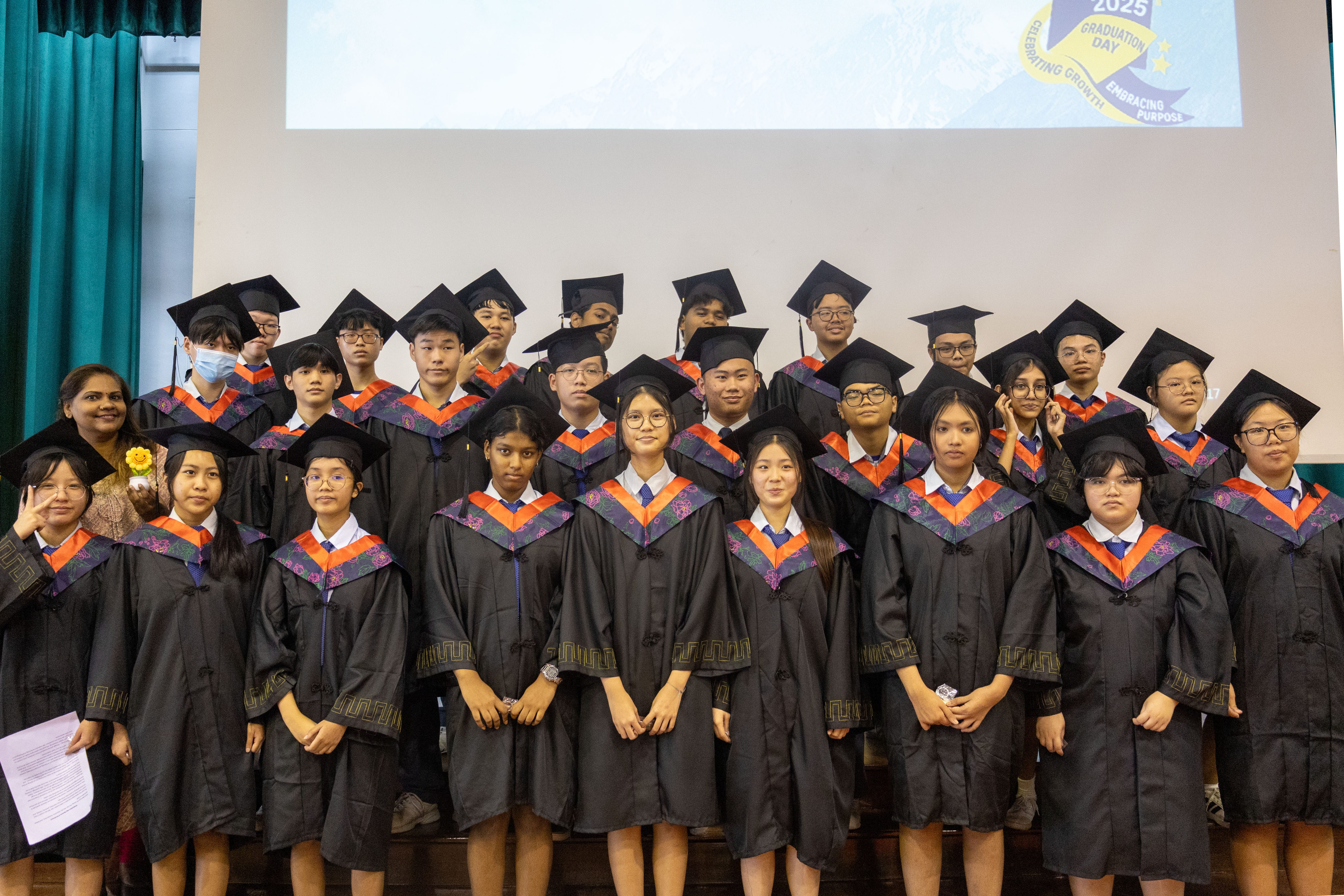 Group of graduates in black gowns and mortarboards posing on a stage beneath a projected 2025 banner