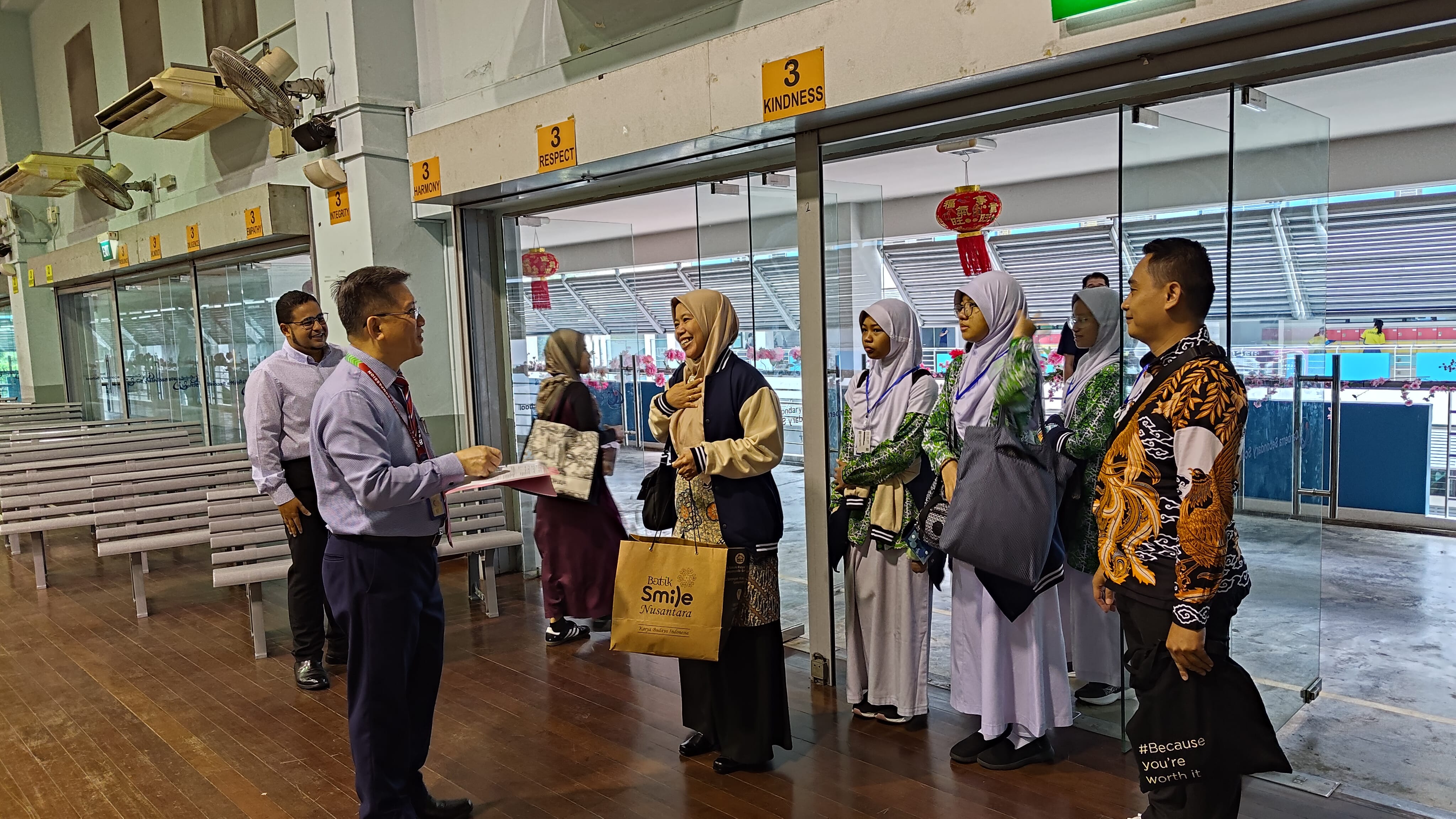 A man in a pink shirt hands documents to a smiling woman in a hijab, with a group of students nearby indoors.