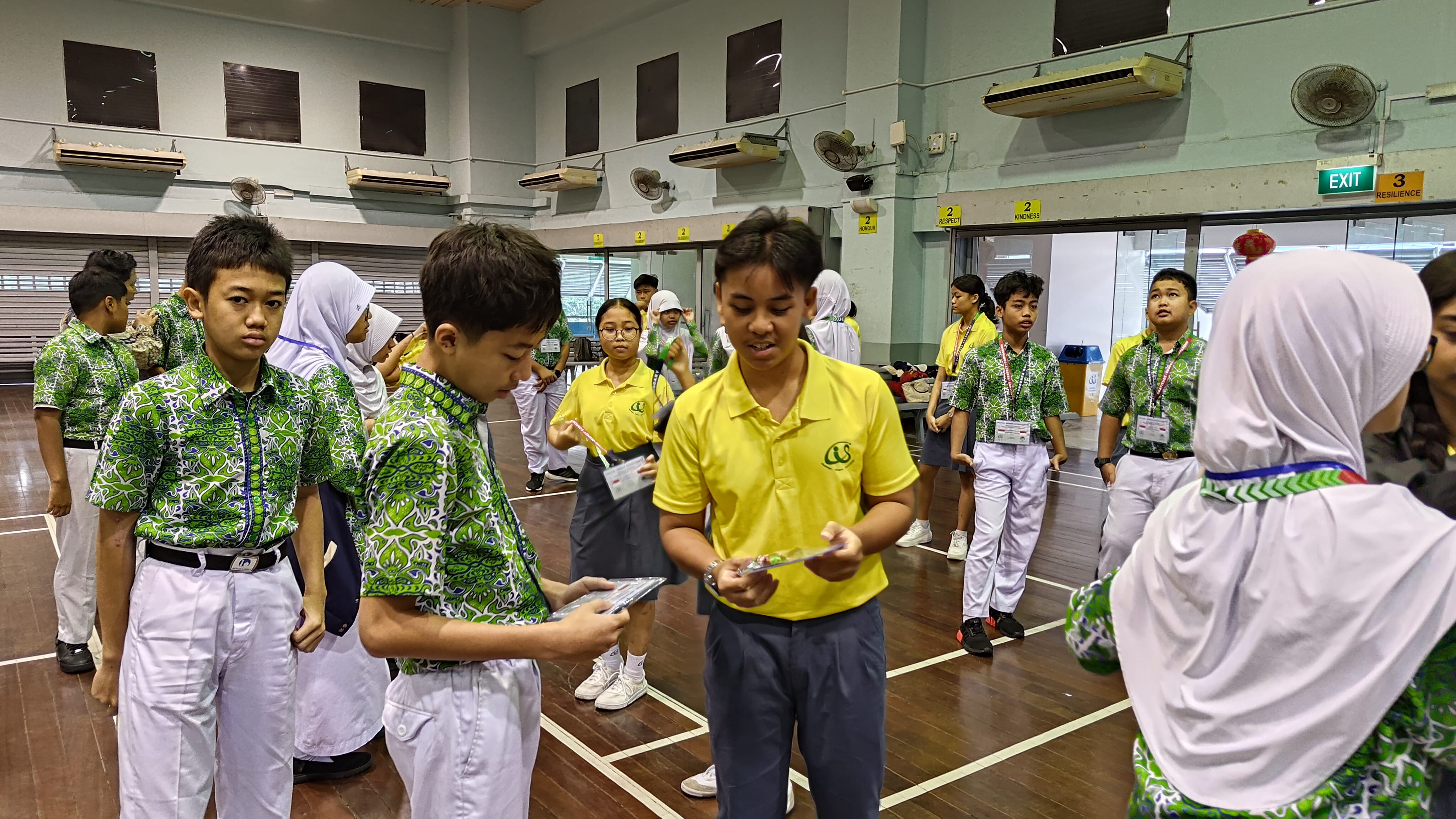 Students in green patterned and yellow uniforms stand in an indoor hall, some holding cards or small items.