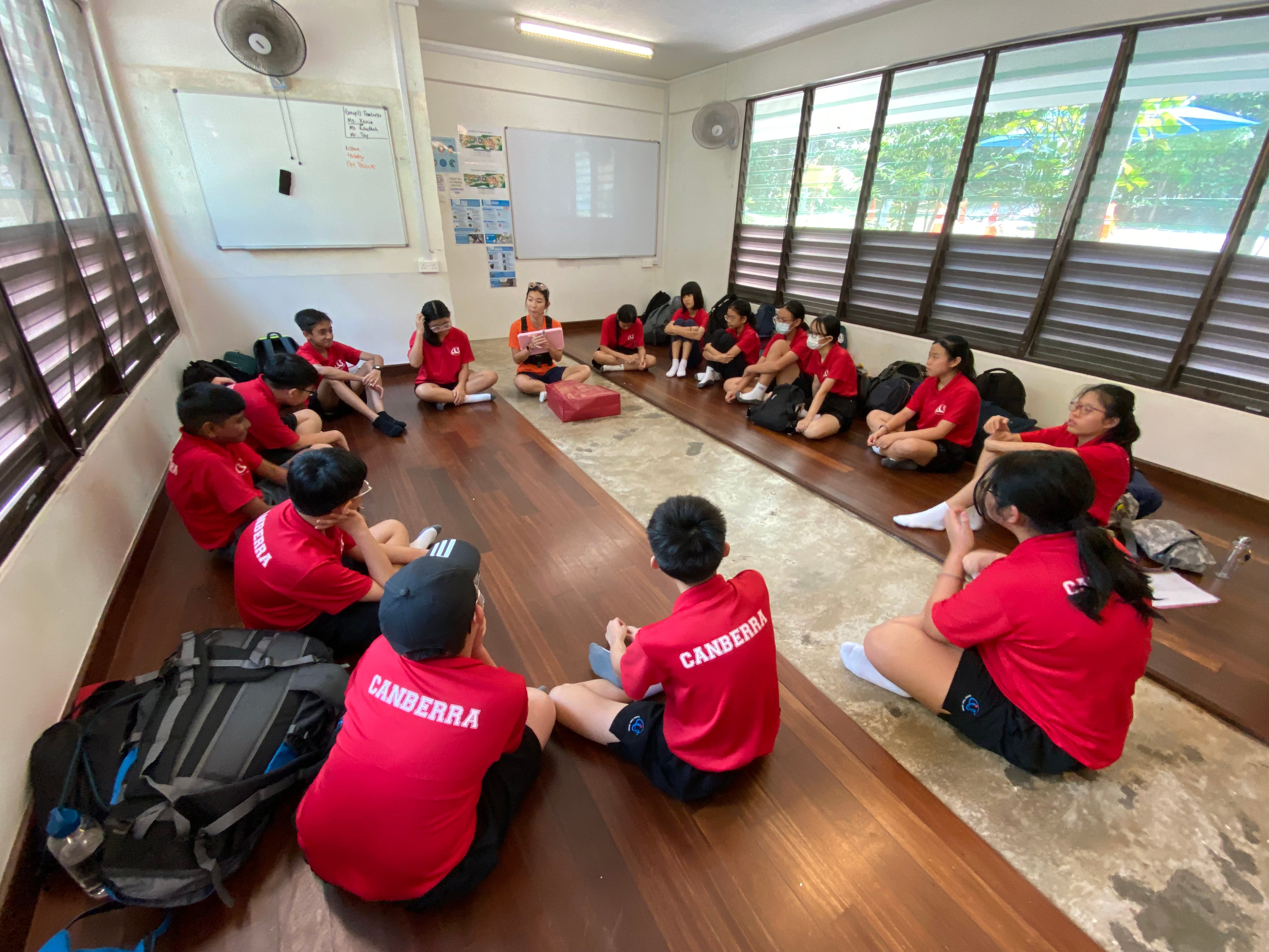Students in red Canberra shirts sit in a circle on a wooden classroom floor during a group discussion.