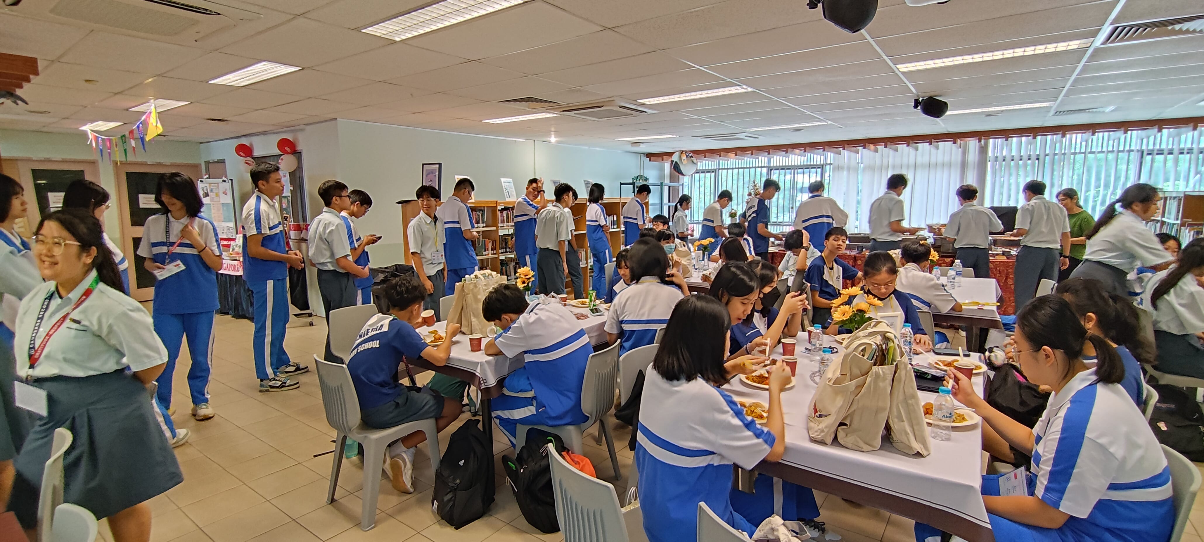 Students in blue and white uniforms eating and socializing at tables in a decorated school hall.