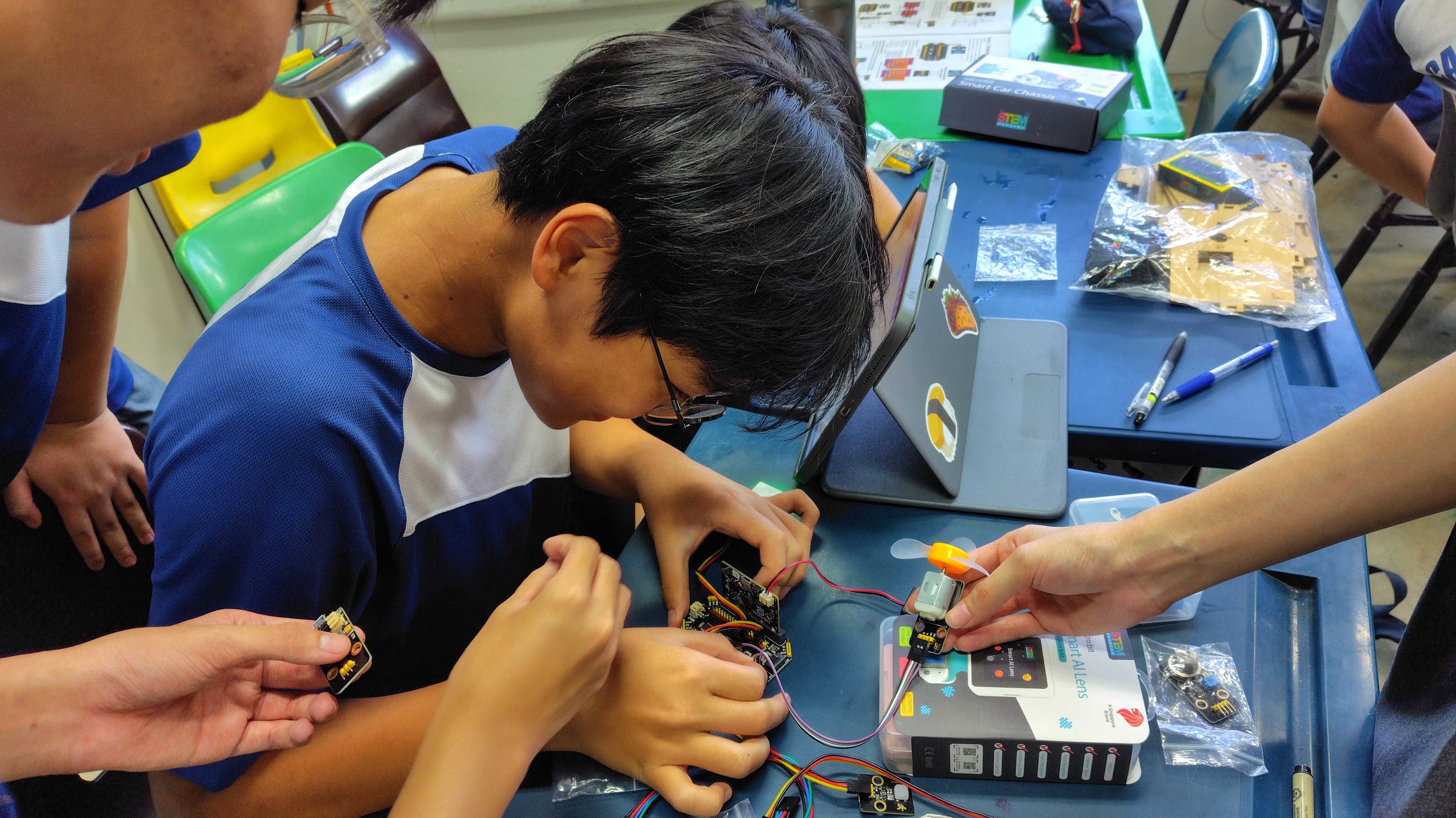A boy in a blue shirt leans close to assemble electronic components with wires on a blue desk during a STEM workshop.