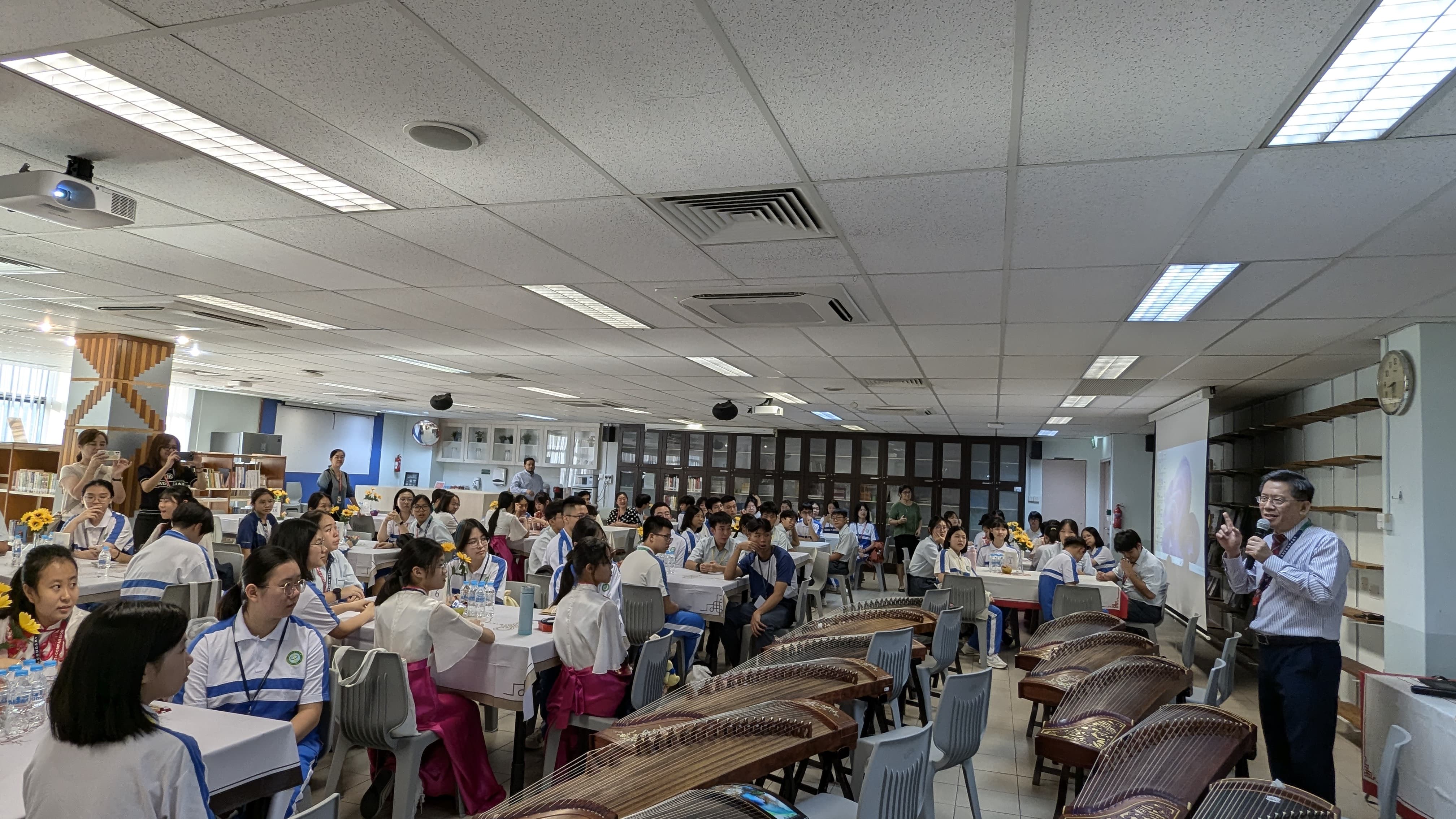 A man speaks into a microphone before a large group of students in uniforms seated at tables, with guzheng instruments nearby.