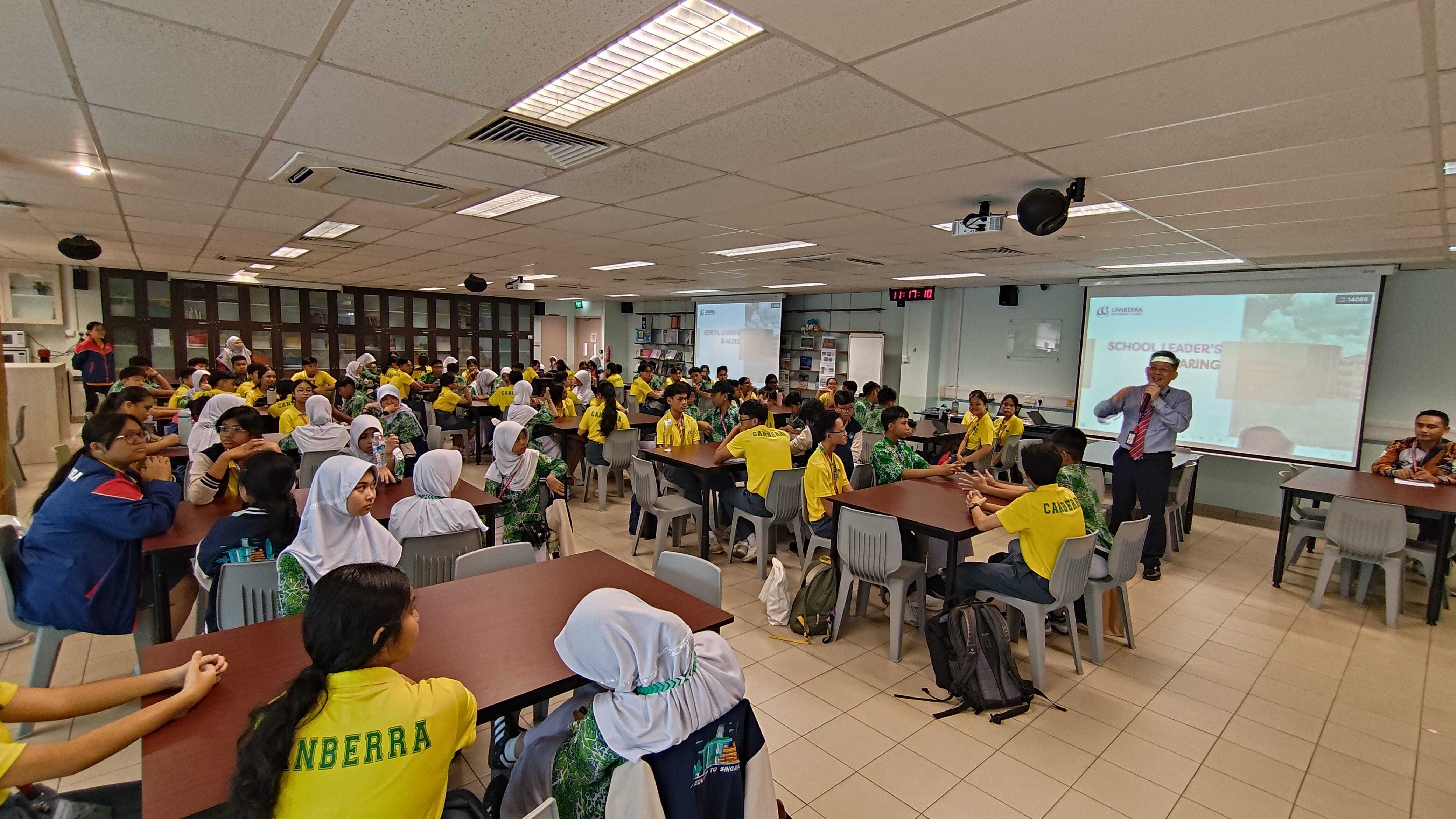 Students in yellow Canberra shirts sit at tables in a school hall while a speaker presents at a projected screen.