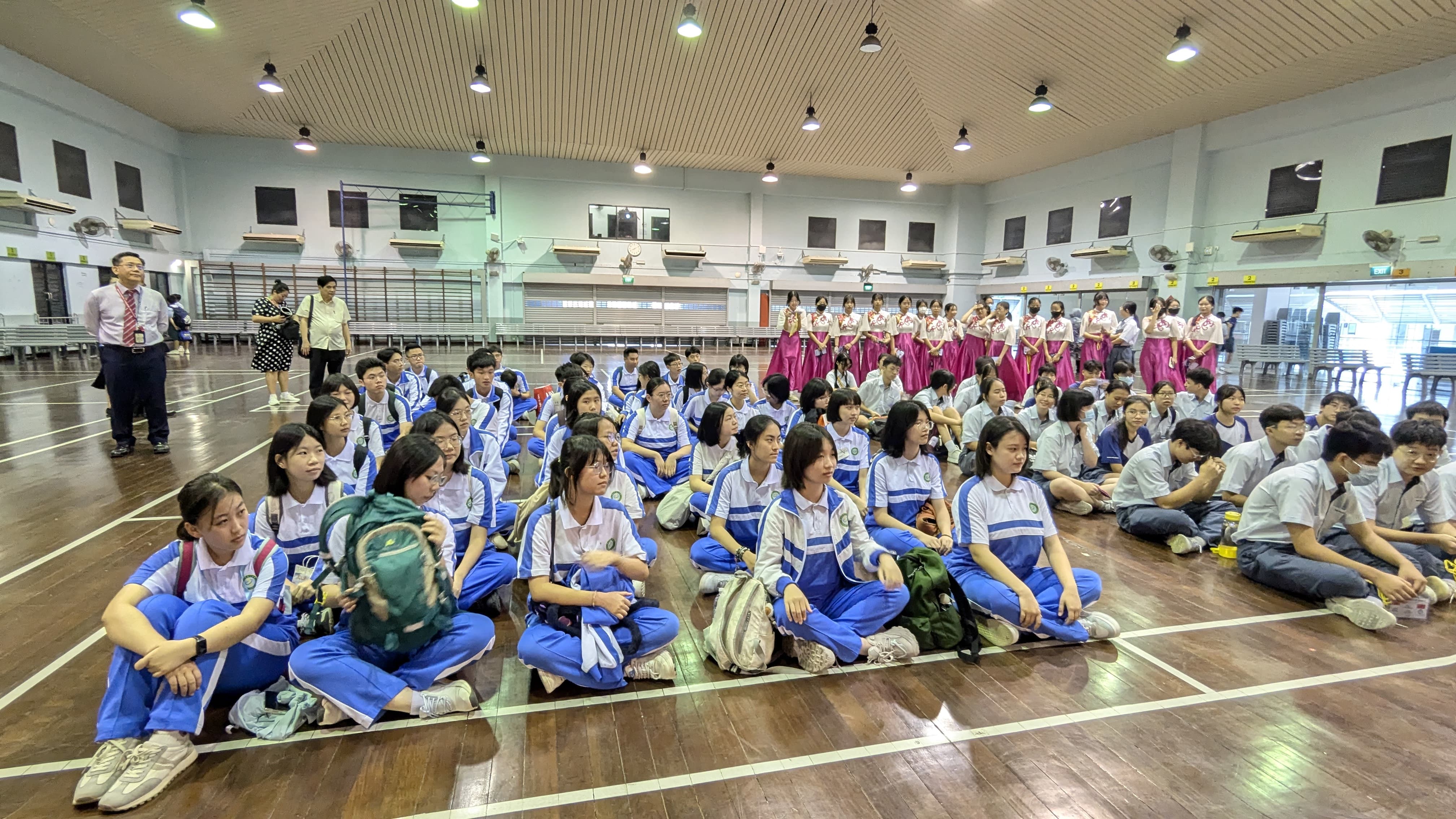 Students in blue uniforms sit on a gym floor facing a group of performers in pink skirts standing at the back.