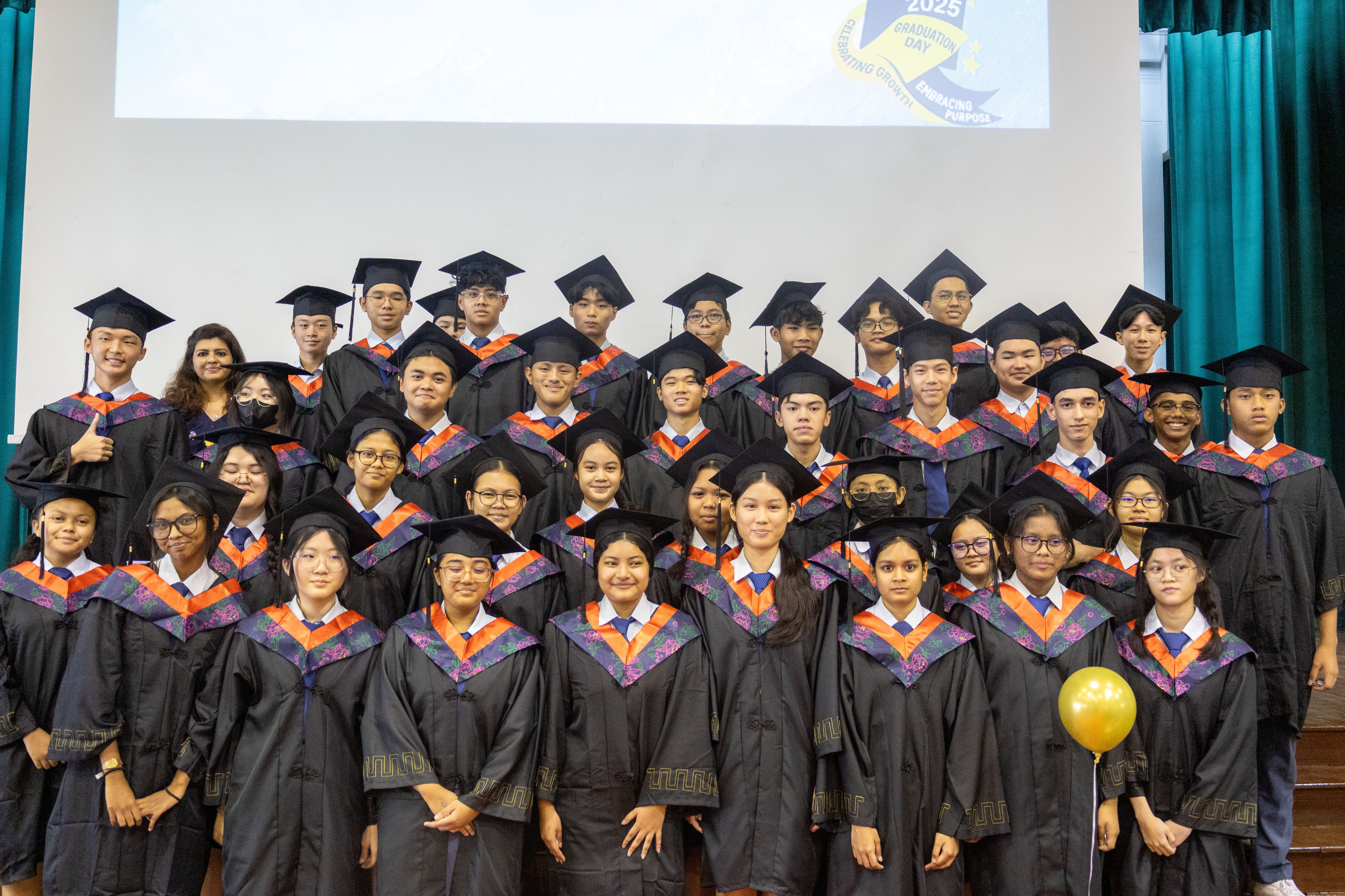 Group of graduates in caps and gowns posing on a stage, with a gold balloon at front right.