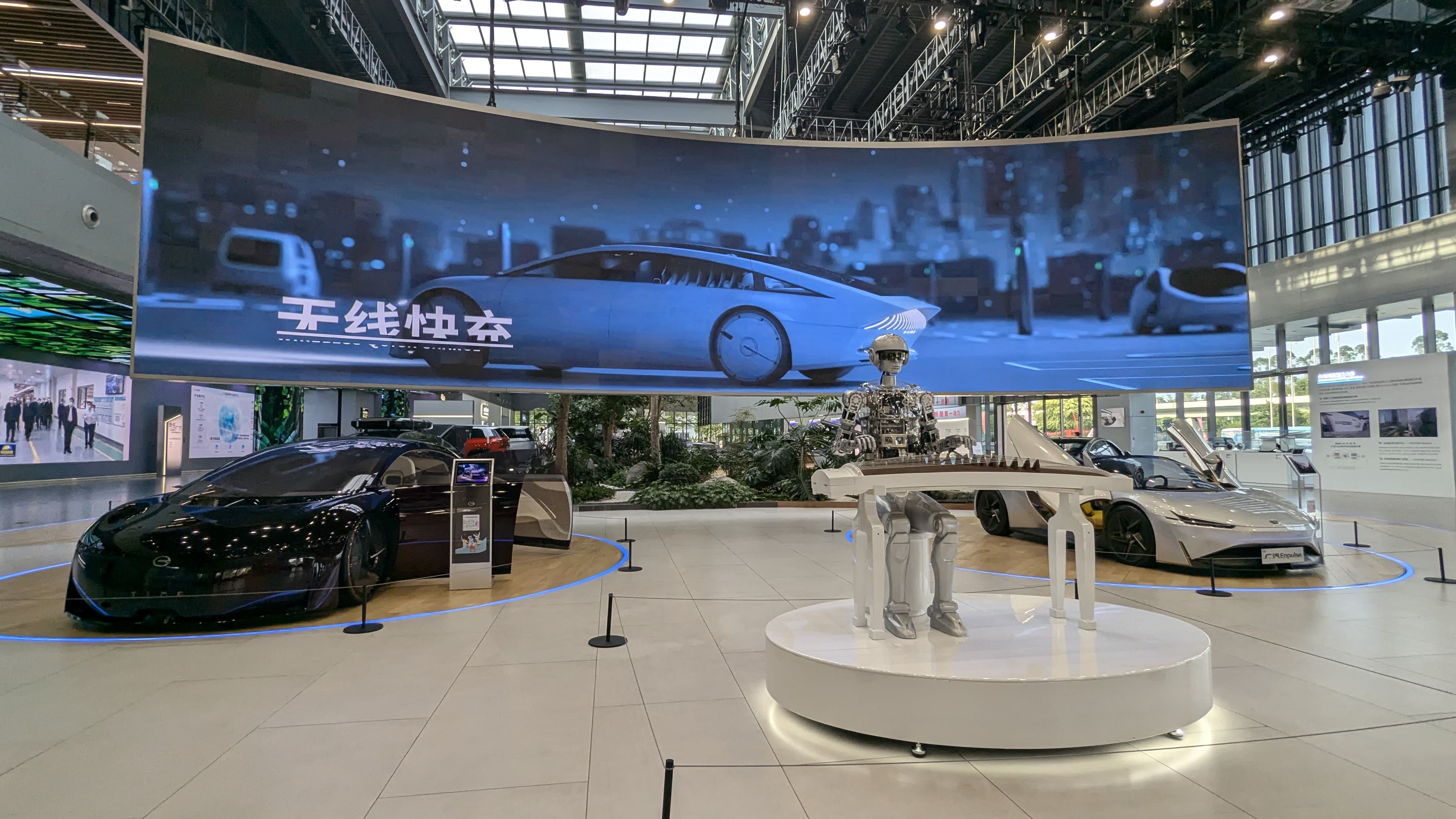 Futuristic car showroom with sleek concept cars, a robot figure on a platform, and a large curved screen with Chinese text.