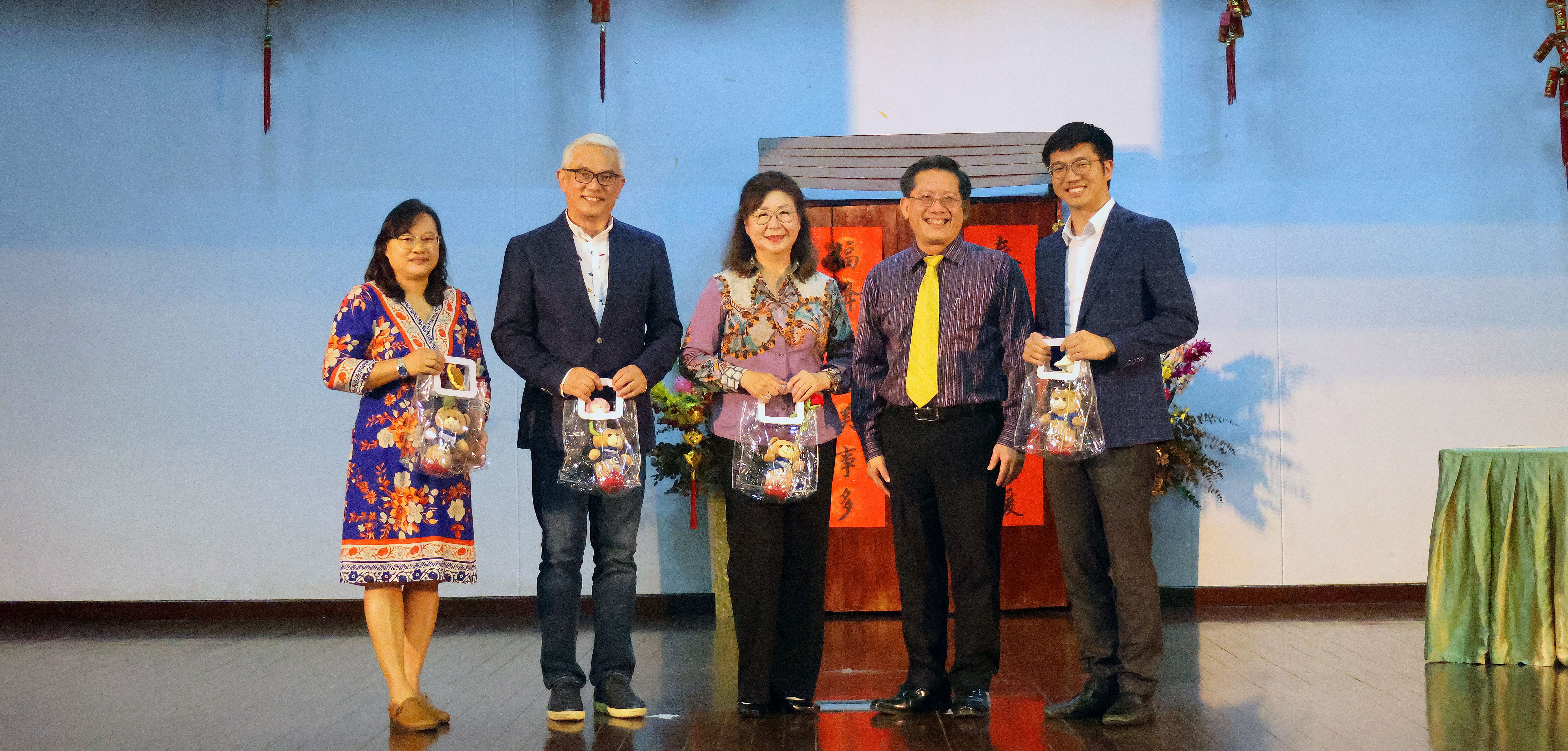 Five adults on a stage smiling, three holding clear gift bags with stuffed toys, podium with red banners behind.