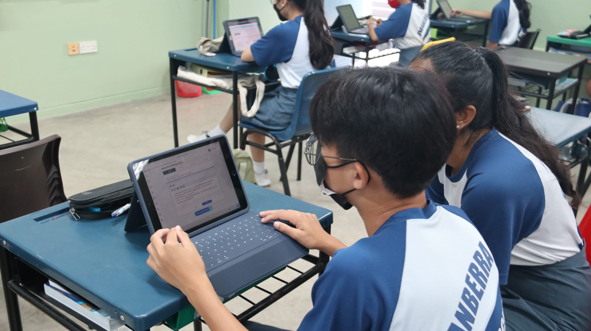 Two students in blue uniforms work on a tablet with a keyboard in a classroom, with more students using devices behind them.