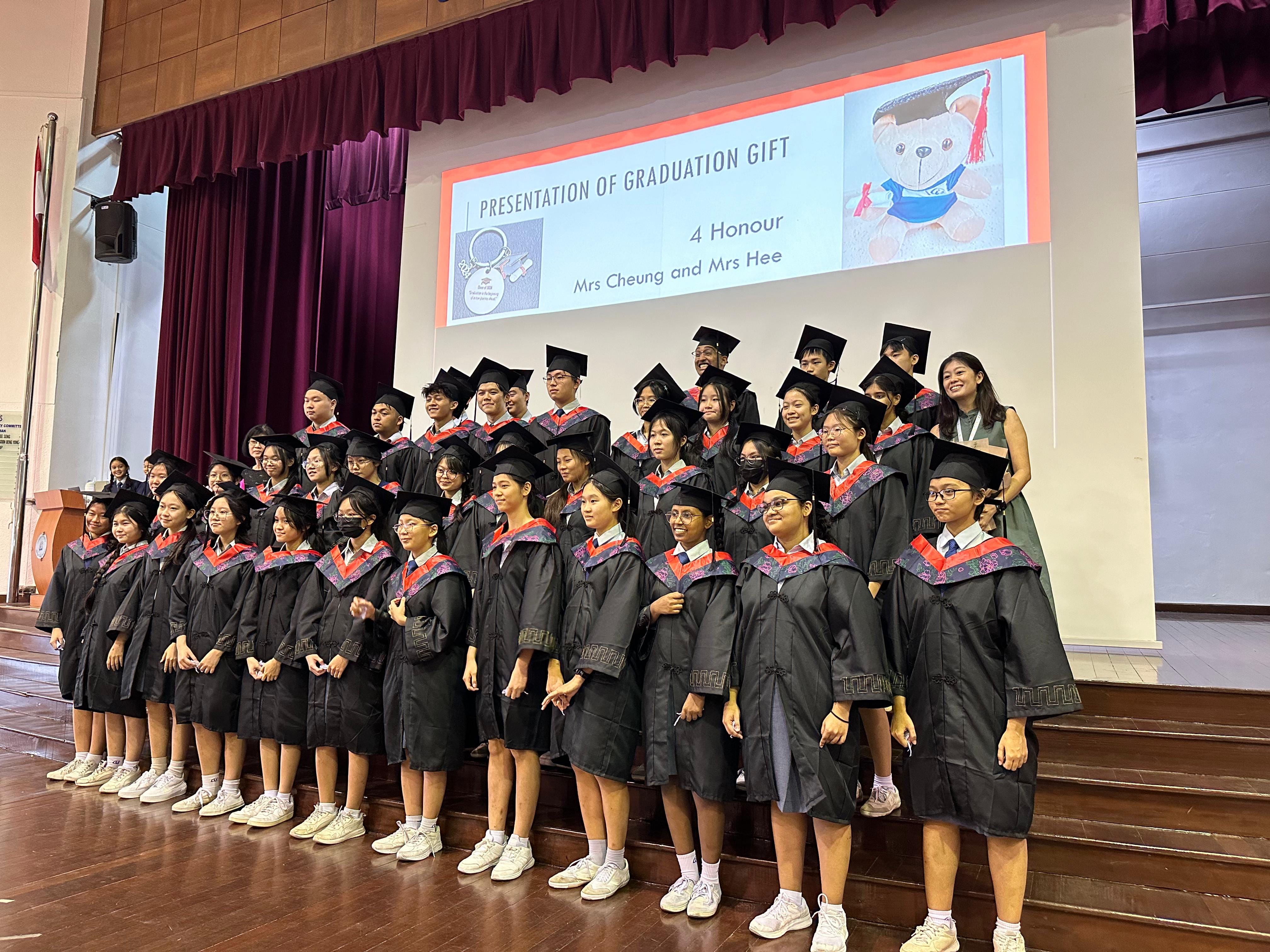 A large group of students in black graduation gowns and caps pose on a stage before a Presentation of Graduation Gift screen.